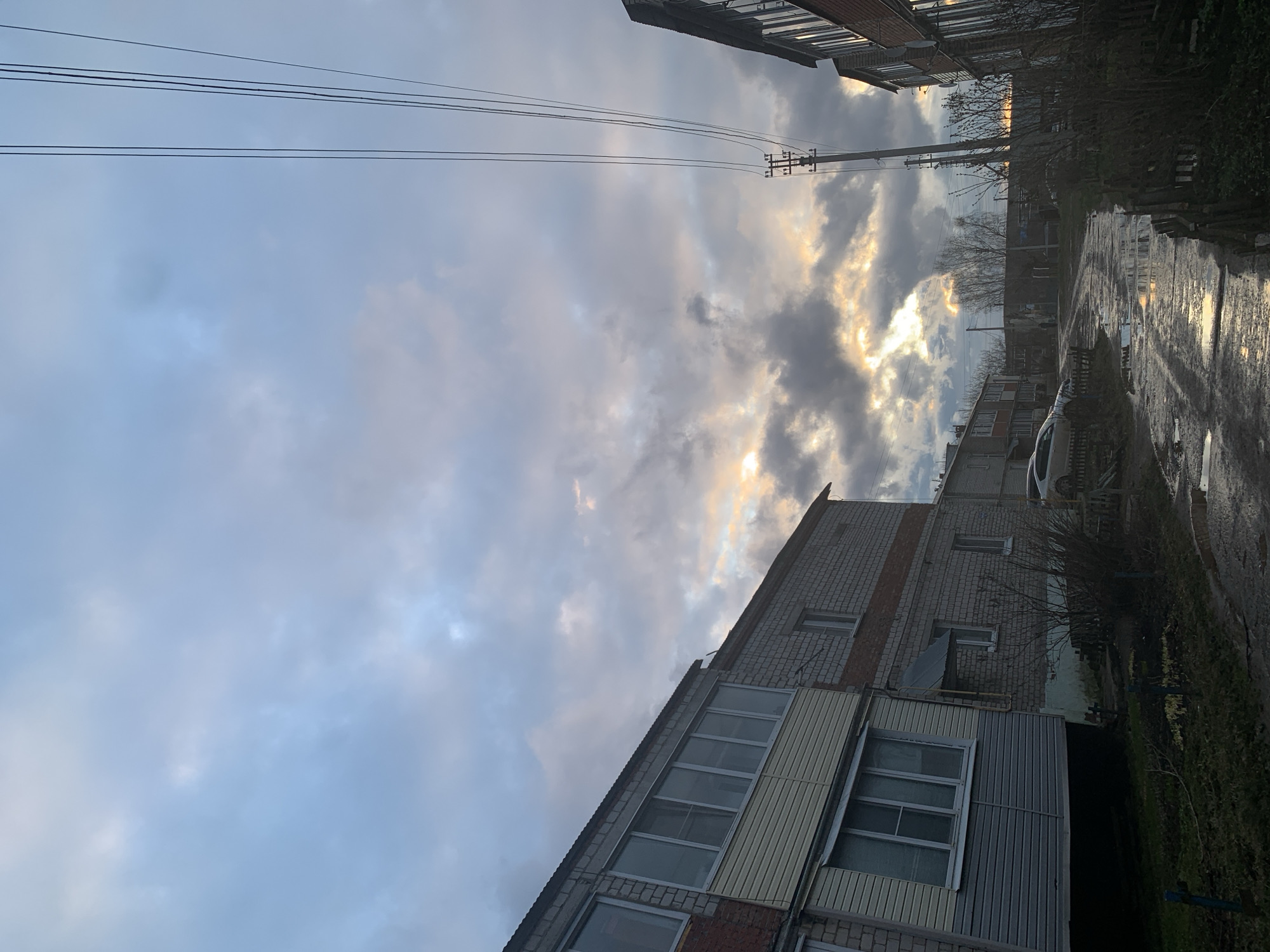 The image captures an outdoor scene in what appears to be a residential area, likely a street or courtyard. The sky is the dominant feature, characterized by a mix of light blue-grey and dark, heavy clouds. Patches of bright, golden sunlight break through the clouds, indicating either sunrise or sunset. Multiple power lines stretch horizontally across the upper portion of the sky, connected to a utility pole on the right side of the frame.
To the left, a multi-story brick building, possibly an apartment block, is partially visible. Another similar building with balconies and light-colored siding on its lower sections is prominent on the right. Between the building and the road, there are some leafless trees and bushes, suggesting either early spring, late autumn, or winter. The ground on the right consists of a wet, unpaved road or path with visible puddles, reflecting the bright sky and suggesting recent rain. A light-colored car is partially visible parked on the right side of the road.
The setting evokes a quiet, somewhat damp atmosphere. The dramatic sky with sunlight piercing through clouds is a notable detail, highlighting the time of day as either early morning or late afternoon. No discernible human activity or legible text is present. The general appearance of the buildings and infrastructure aligns with typical residential areas in a Russian town or city, consistent with the given location context of Фоки, Russia.