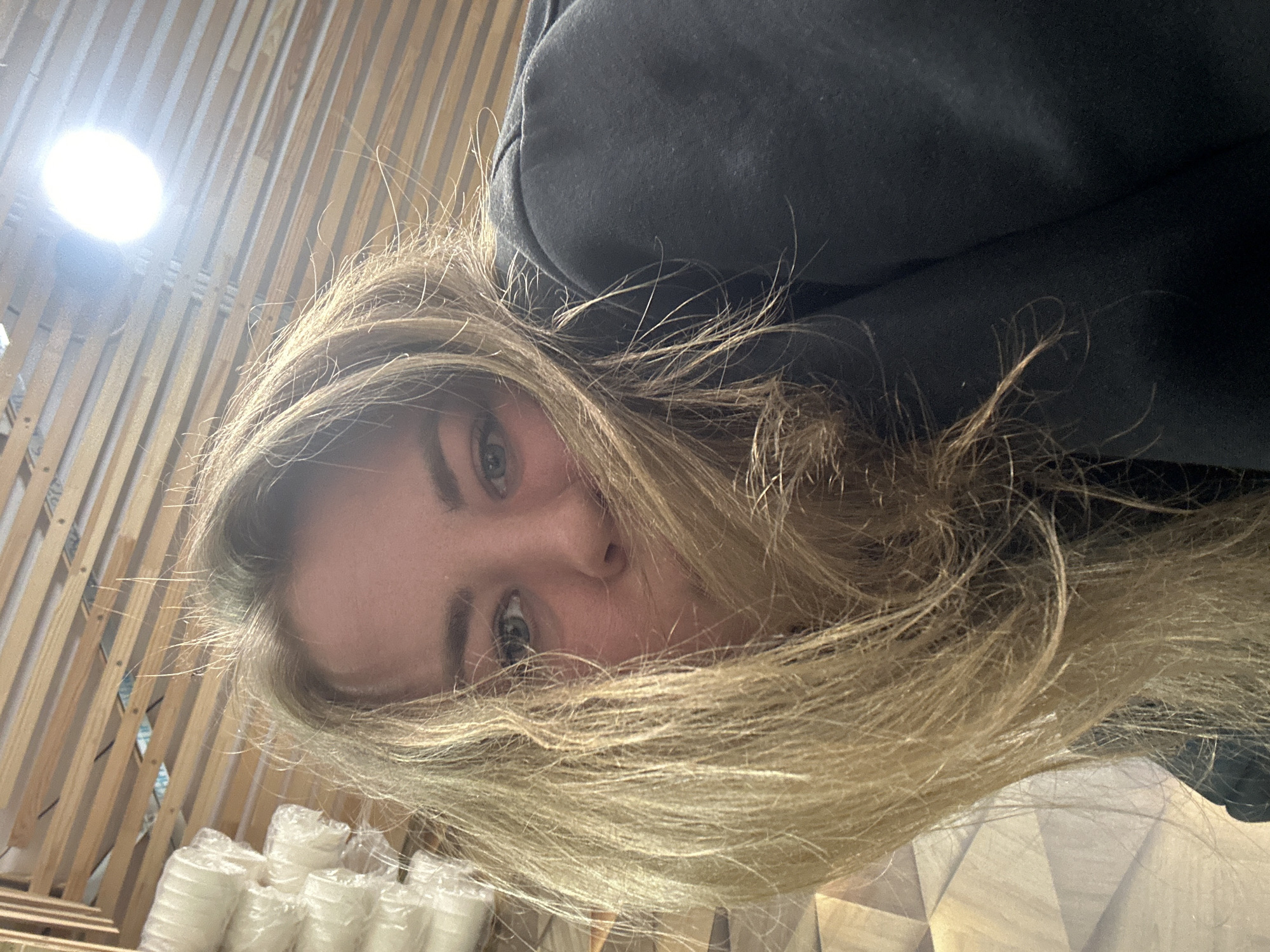 A close-up, angled portrait features a young woman with light-colored eyes, possibly blue or green, gazing directly at the camera. Her long, blonde hair is prominent, framing her face and partially covering parts of her forehead and cheeks. She is wearing a dark, likely black, hooded garment such as a sweatshirt or jacket.
The setting appears to be indoors. In the upper left corner, a bright, circular light source causes a lens flare. The background behind her left side reveals a patterned wall or partition made of vertical wooden slats. On the lower left, below the wooden structure, are multiple stacks of white, cylindrical objects, individually wrapped in clear plastic, which resemble disposable cups or small bowls. To the lower right, another surface with a geometric, possibly wooden or tiled, pattern is visible. The overall impression is a casual, intimate shot taken in a modern indoor environment. There is no visible text.