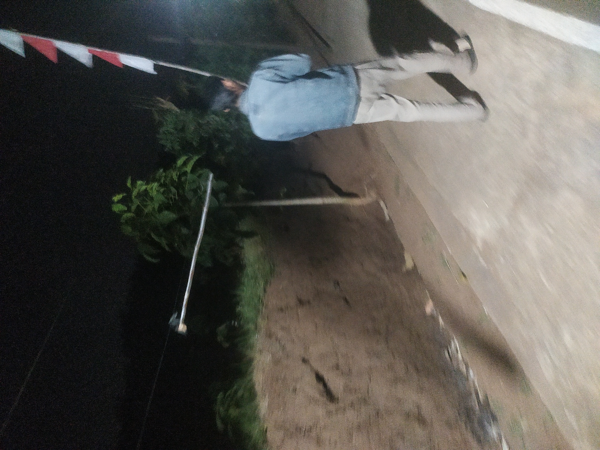 A person stands on a paved path at night, holding a pole with several red and white flags attached. The person is wearing a light blue denim jacket and light-colored pants. They are facing away from the camera. To the left of the path, there is a sloped dirt embankment with some greenery. In the background, there are trees and a dark sky. The scene appears to be outdoors, possibly in a rural or semi-rural area. The location of Lopait, Indonesia is noted.