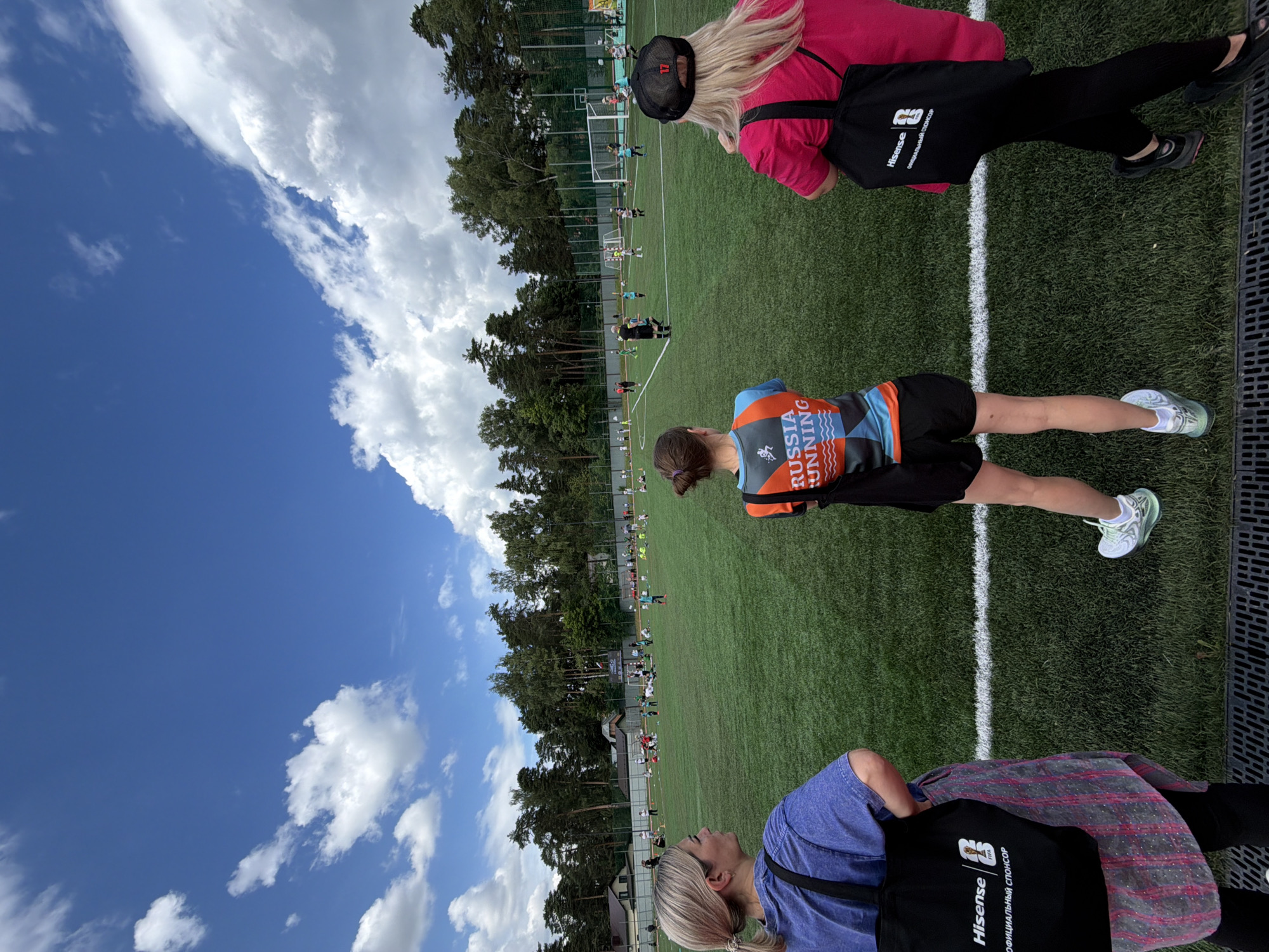 The image shows an outdoor artificial turf sports field, likely in the city of Bykovo, Russia, as indicated by the context. The field is lined with white markings, and there are several children and adults playing on it. The weather appears to be fair, with a blue sky and scattered white clouds. In the foreground, three individuals are visible from behind, observing the activity on the field. One person on the left is wearing a blue t-shirt and a black tote bag with "Hisense" and "FIFA" logos. The person in the middle is wearing an orange and blue athletic top that reads "RUSSIA RUNNING" and black shorts. The person on the right is wearing a pink t-shirt and a black cap with a white number "17". The background features trees and some residential buildings, suggesting a suburban or park-like setting.