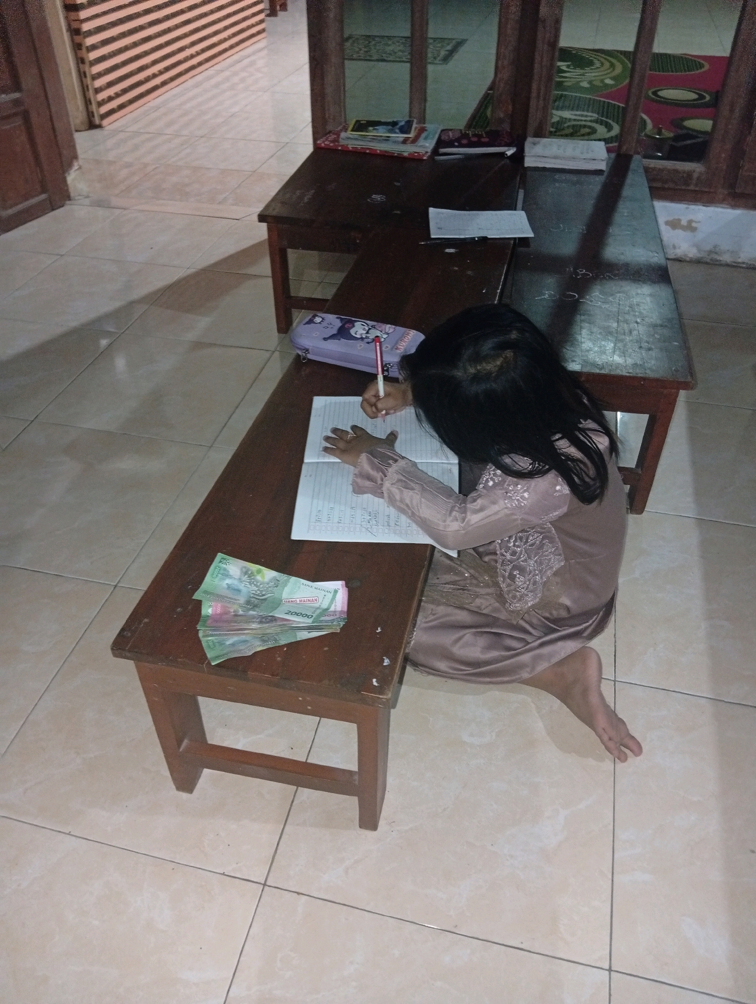 The image depicts a young girl sitting on the floor in front of a wooden desk, engaged in writing in a notebook. She is wearing a beige dress and her back is turned towards the camera. On the desk, there is a pencil case, the notebook, and several Indonesian Rupiah banknotes. The environment appears to be indoors, with tiled flooring and wooden furniture, suggesting it might be a classroom or a home study area. The lighting is dim, creating shadows on the floor and furniture. The desk itself is made of dark wood and has a simple, rustic design. The overall scene suggests a moment of focused study or homework.