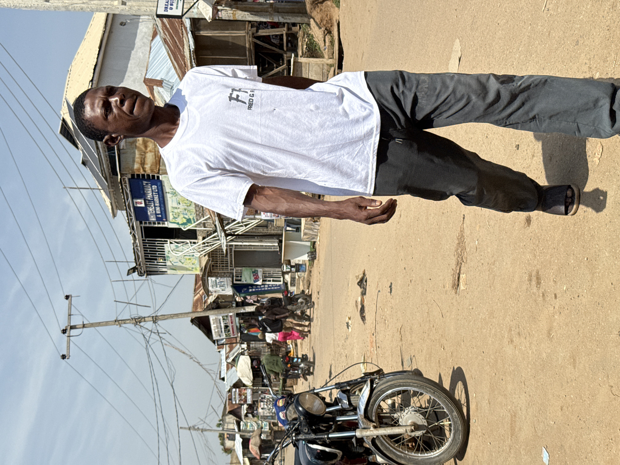 A man walks down a dusty, unpaved street in Abuja, Nigeria. He is wearing a white t-shirt with a logo that reads "FRED & G" and dark grey pants. The street is lined with small shops and stalls, some with corrugated metal roofs. A motorcycle is parked on the left side of the image. In the background, there are power lines crisscrossing overhead and several buildings with various signs, including one for "WHITEHEART PHARMACY & MINI-MARKET LTD." The sky is clear and blue. Several other people are visible in the background, some walking and others on motorcycles.