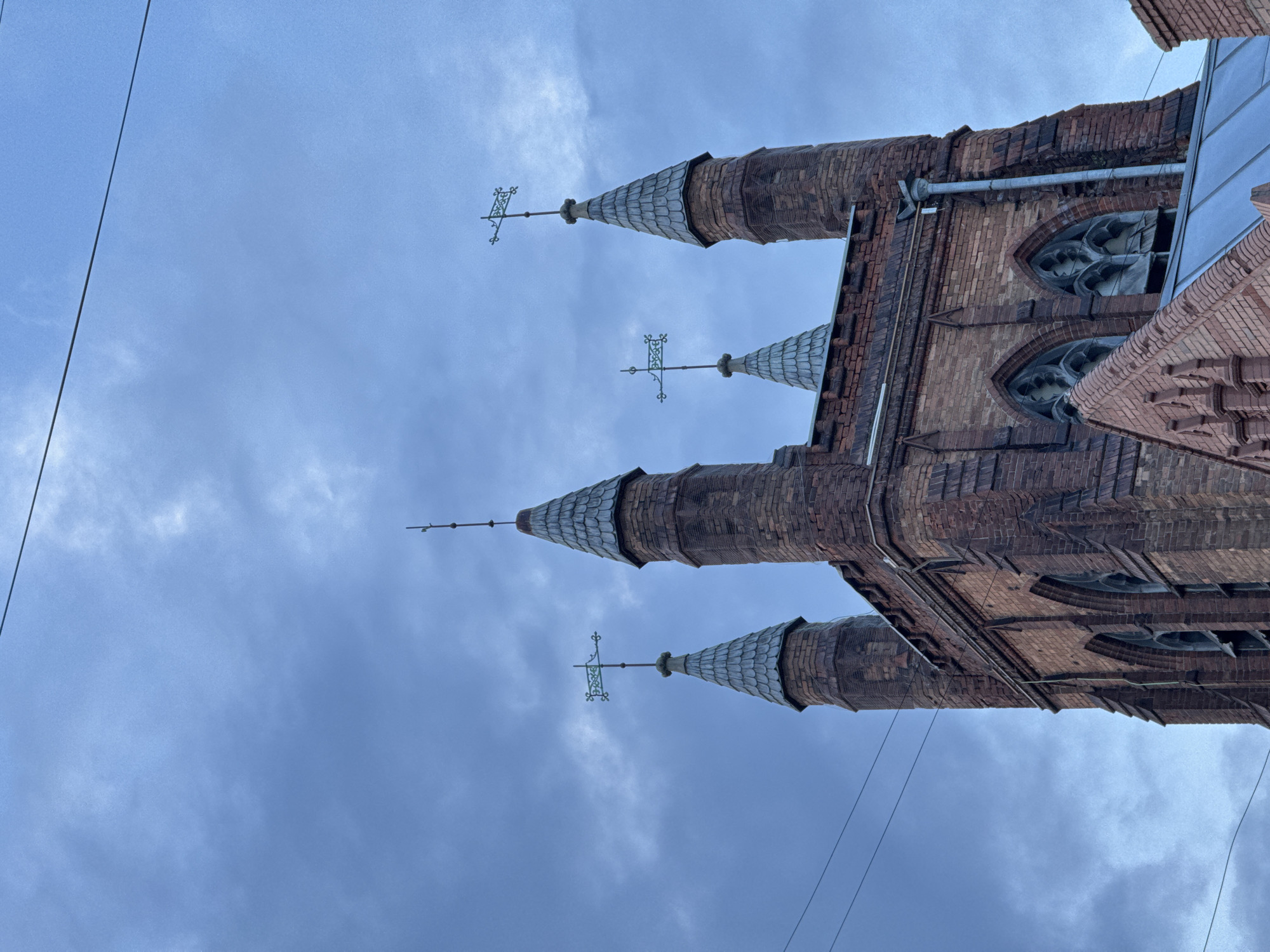 The image depicts a low-angle view of the Evangelical Lutheran Cathedral of Saint Peter and Paul in Moscow, Russia. The building is made of red brick and features multiple spires topped with weather vanes. The sky is overcast and cloudy. There are power lines visible in the lower left and right corners of the image. The cathedral's architectural details, including the Gothic-style windows and brickwork, are visible.