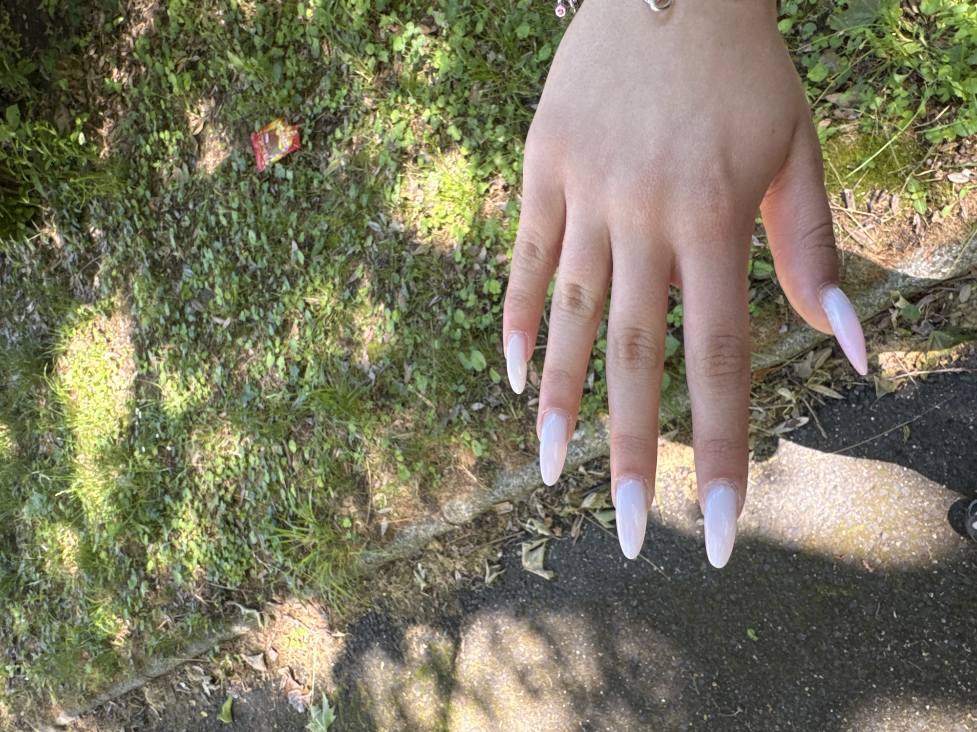 A pale-skinned hand with long, almond-shaped, light-colored nails is positioned over a mixed environment of grass and concrete. A bracelet adorns the wrist of the hand. A small, colorful candy wrapper is visible amidst the grass. The overall environment is dappled with sunlight.