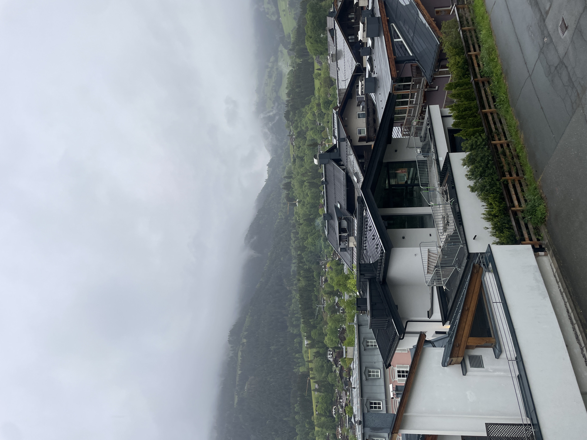 A high-angle view shows a landscape including trees and buildings under cloudy sky. The right side of the image contains multi-story buildings with black roofs and white or gray exterior walls. Some of the buildings feature balconies with metal railings. A wooden fence and a strip of green foliage run along the edge of a paved street or walkway in front of the buildings. The left side of the image shows a forested hillside with trees and some open grassy areas. Mist or fog hangs in the air, partially obscuring the view of the hillside. The city of Unknown, Unknown, is not determinable.