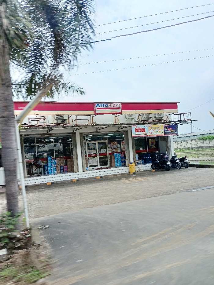 The image captures an Alfamart convenience store in a semi-urban or rural setting. The main subject is the single-story store building with a prominent red and white facade and the "Alfamart" logo. A tall palm tree stands on the left, partially obscuring the view.

In front of the store, there's a paved area serving as a parking space where three motorcycles are parked. A single person is visible sitting on a bench near the left entrance of the store. The store's interior is visible through large glass windows, showing various products, including stacks of bottled water or similar items. A yellow trash bin is placed near the right entrance. Overhead, utility lines stretch across the cloudy sky. In the background to the right, a low wall and what appears to be an open field or agricultural land are visible.

The scene suggests a typical daytime setting under an overcast sky, indicating mild weather without direct sunlight. The store appears to be open for business, with the parked motorcycles implying customer activity. The presence of Alfamart confirms the location as Indonesia, aligning with the Ngoro context.

The most prominent visible text is "Alfamart" on the store's sign. A promotional banner hanging outside the store on the right displays "20.000," likely indicating a price or offer. Other text inside the store and on the banner is too small or blurry to be legible.