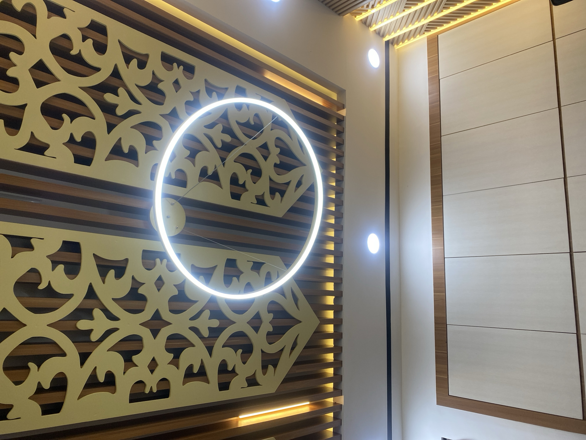 The image shows a modern ceiling with intricate golden geometric patterns overlaying wooden slats. A circular white light fixture hangs centrally, illuminating the patterned ceiling and the space below. The ceiling is framed by recessed warm-toned lighting. Below the ceiling, a section of a wall is visible, featuring light-colored cabinet doors with subtle wood trim. Another wall on the right side of the image has vertical wooden slats with integrated yellow lighting. The overall aesthetic is contemporary and sophisticated. The location is specified as Banjarbaru, Indonesia, suggesting this is likely an interior shot of a residential or commercial space within that city. No people or specific actions are depicted.