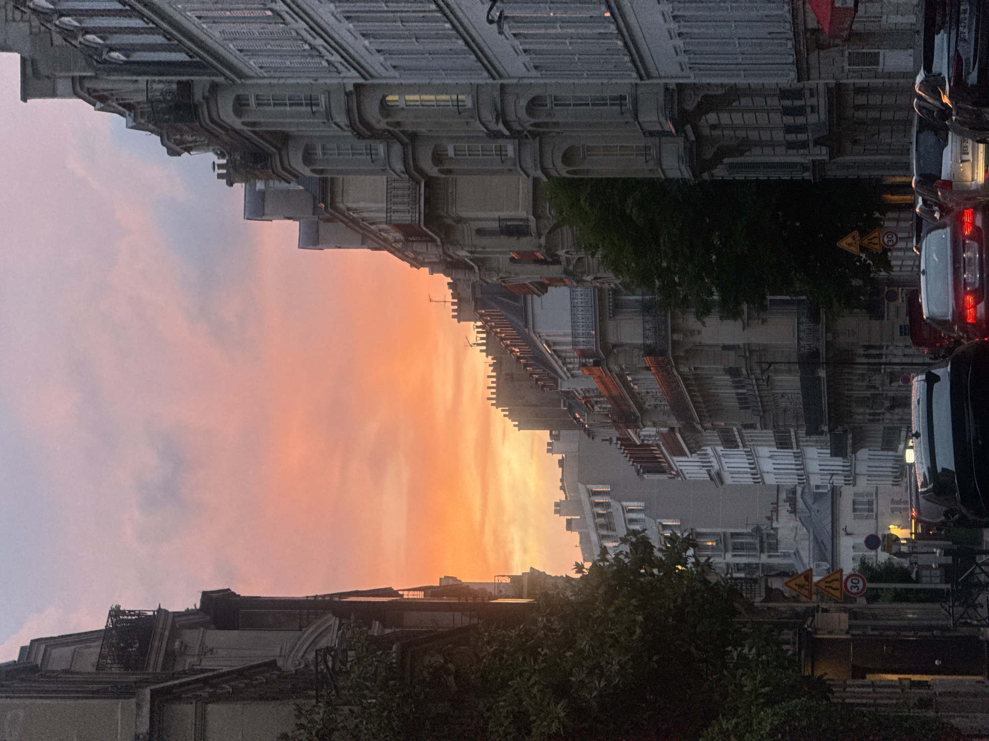 The image captures a street scene in Paris, France, at twilight. The sky displays a gradient of colors from pink and light gray near the horizon to a fiery orange closer to the buildings, indicating the setting sun. Tall, ornate Parisian buildings line both sides of the street, showcasing classic architectural details like windows, balconies, and cornices. A row of parked cars is visible on the right side of the street. Street signs are also present, including those indicating speed limits. There are trees whose branches extend in front of the buildings. The perspective suggests the photo was taken from street level, looking down the road.