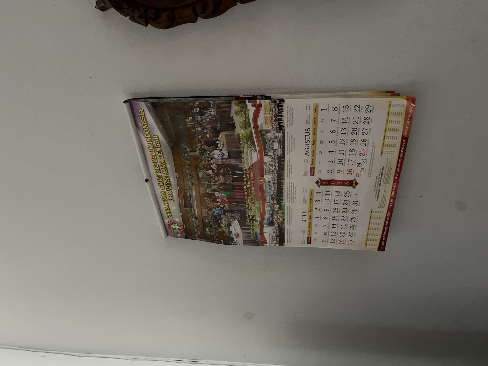 The image shows a calendar hanging on a light gray wall. The calendar is for the year 2026 and displays the months of July and August. It features an image at the top depicting a group of people in formal attire, likely a gathering or event. Below the image, the calendar lists dates, days of the week, and some additional text in Indonesian. The calendar is partially obscured by shadows and the angle of the shot. The wall appears to be an interior surface, with no specific architectural details indicative of a particular location within Glagah Kulon, Indonesia, beyond it being an indoor setting. There are no people or discernible activities captured within the scene other than the calendar itself.