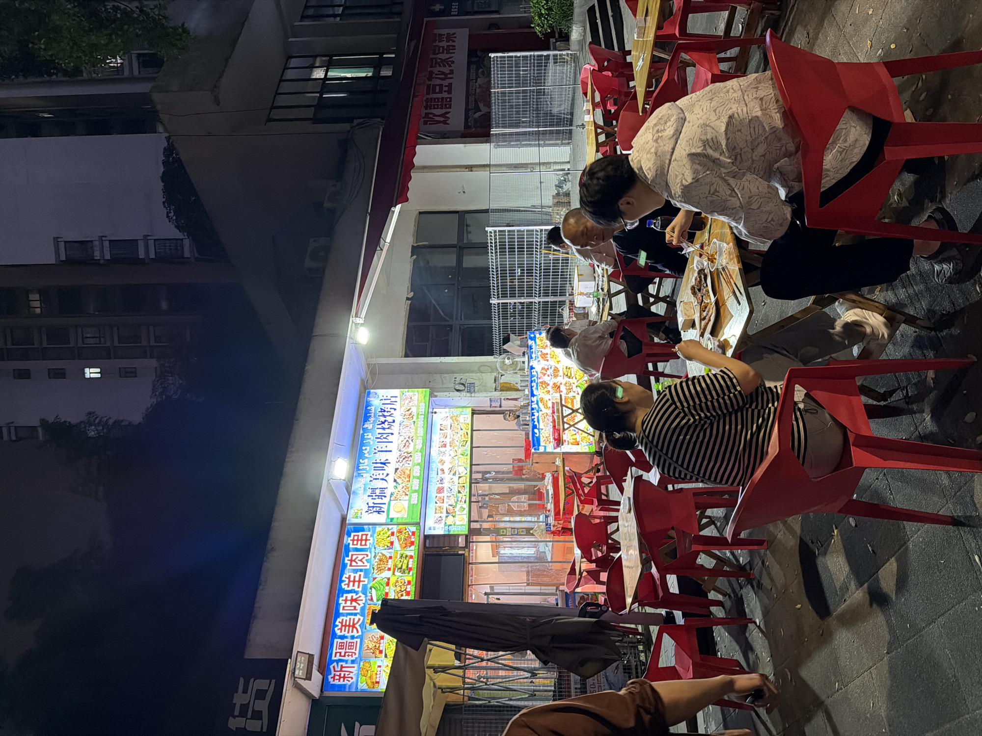 A nighttime scene in the Chongqing High-tech Zone, China, depicts an outdoor restaurant setting. Several people are seated at tables, engaged in eating. The red plastic chairs are a dominant feature of the scene. Above the patrons, bright signage with Chinese characters is visible, indicating the name of the restaurant and dishes, including Xinjiang-style lamb skewers. The restaurant's façade is illuminated, contrasting with the dimly lit street. Some structures are covered with scaffolding and tarps.