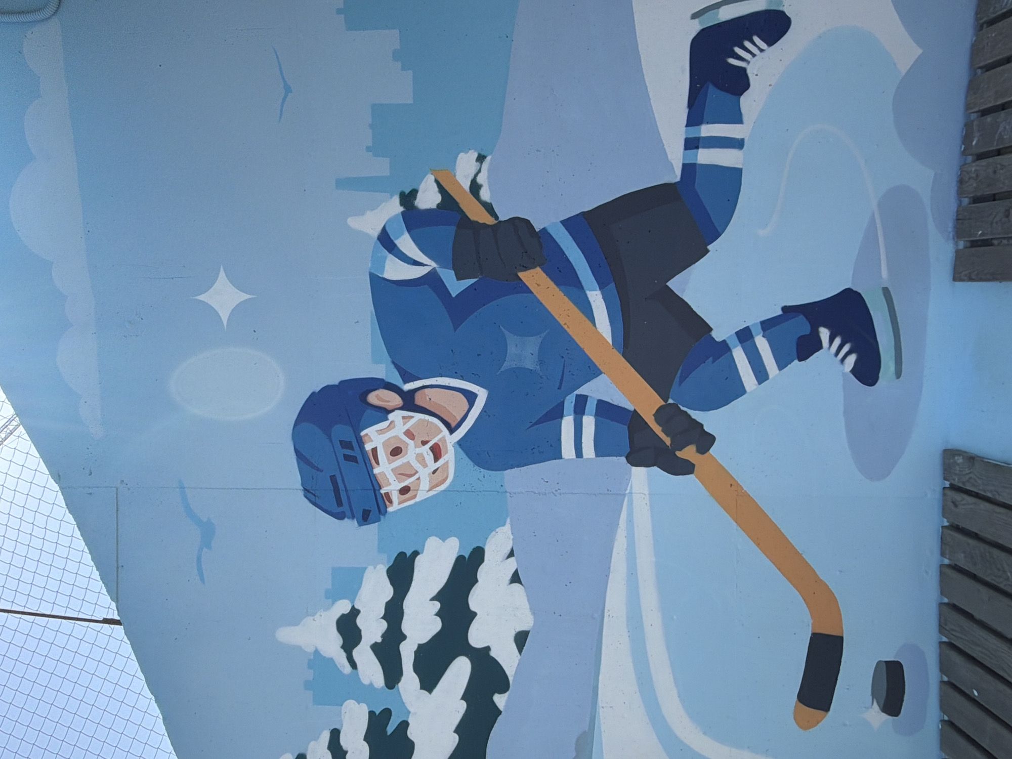 This is a mural on a concrete wall depicting a hockey player in a blue uniform with white stripes. The player is wearing a blue helmet and holding a hockey stick, with a puck visible on the ice in front of them. The background shows a winter scene with snow-covered trees, a city skyline in silhouette, and birds in a pale blue sky. There are also faint sun or cloud shapes in the sky. Wooden benches are visible at the bottom of the mural. The mural is located outdoors, near a chain-link fence. No specific location in Yekaterinburg, Russia is indicated in the image.