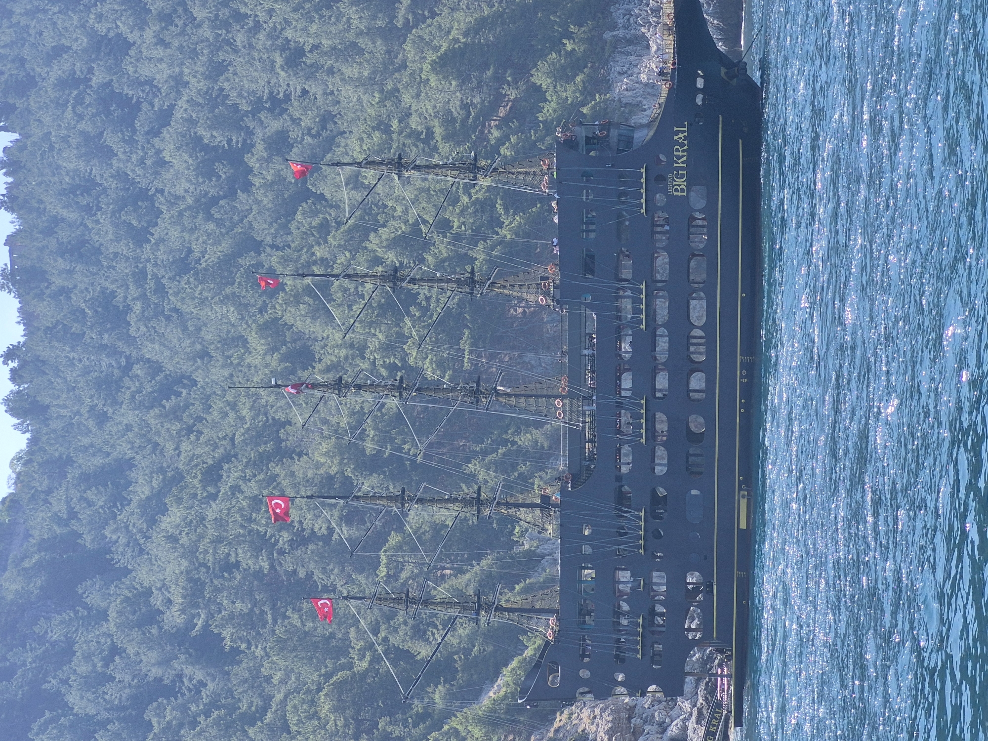 A large, black, pirate-themed tourist ship is docked in Alanya, Turkey. The ship is named "Big Kral Legend." It has multiple masts flying Turkish flags. The ship is docked against a rocky coastline covered in green trees. The water is light blue and glistens in the sunlight.