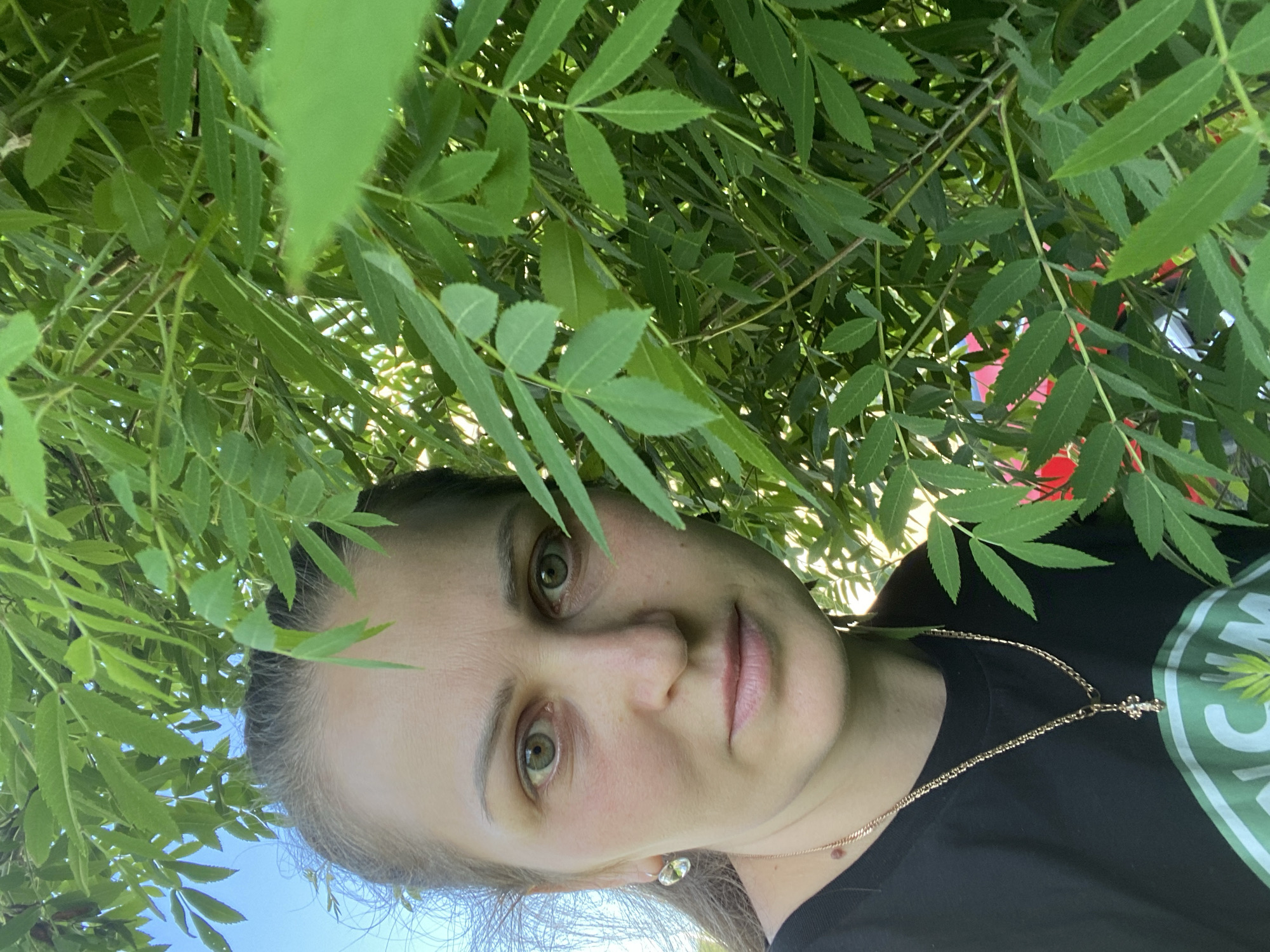 A woman is facing the camera, with green leaves framing her face. She is wearing a black t-shirt and a gold necklace. The photo appears to be taken outdoors, with some blue sky visible. There's no information available to confirm the specific location is Марковский, Russia.
