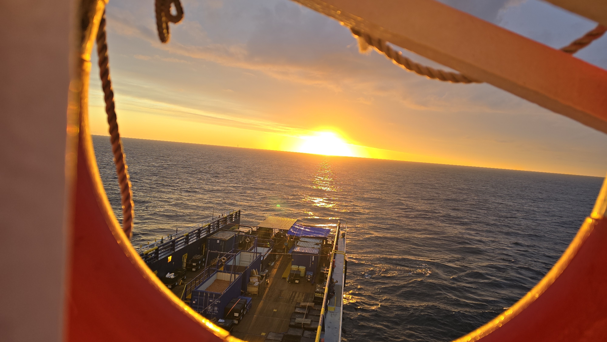 The image captures a sunset over the ocean, as viewed through a framed opening, possibly from a vessel. The sun is low on the horizon, casting a golden light across the sky and reflecting on the water's surface. Below the horizon line, a section of a ship's deck is visible, containing cargo and equipment. The location is the open ocean. The city and location are not known.