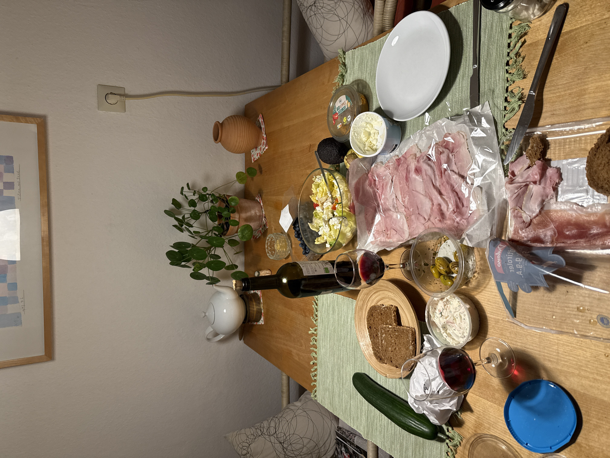 An overhead view of a wooden table laden with a meal, set in an indoor environment that appears to be a dining room in Munich, Germany, based on the presence of a typical European electrical outlet on the wall. The table is covered with a green placemat with fringe. In the center, a bottle of red wine stands next to a glass filled with red wine. A bowl of salad, containing lettuce, tomatoes, and cheese, is also present. Slices of ham are neatly arranged on a plastic package. To the left, a cucumber and two slices of dark bread on a wooden plate are visible. There are also several small containers with dips and olives. A white teapot sits on a decorative coaster in the background, alongside a potted plant and a terracotta vase. The wall behind the table features a framed piece of art and an electrical socket with a cord plugged in. The overall scene depicts a prepared meal, suggesting a dining setting.