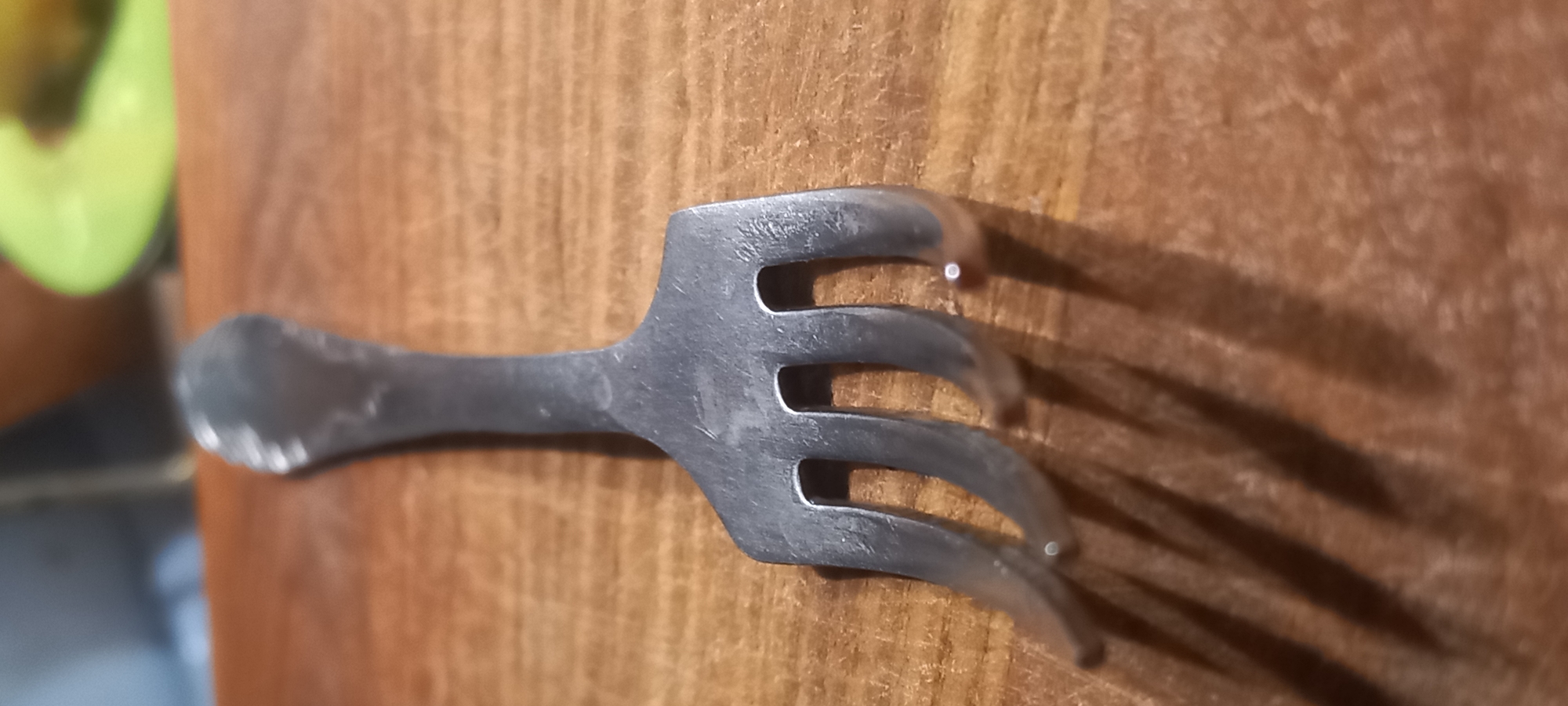 The image shows a metal fork lying on a wooden surface. The fork is positioned at an angle, with its tines pointing downwards and slightly to the left. The tines are curved and appear to be made of silver or stainless steel. The wooden surface is a light brown color and has a visible grain pattern. There are shadows cast by the fork onto the surface. In the background, out of focus, is a blur of colors which might suggest other objects or a background environment, possibly including a lime green object. No specific details about Kryvyi Rih, Ukraine are present in the image.