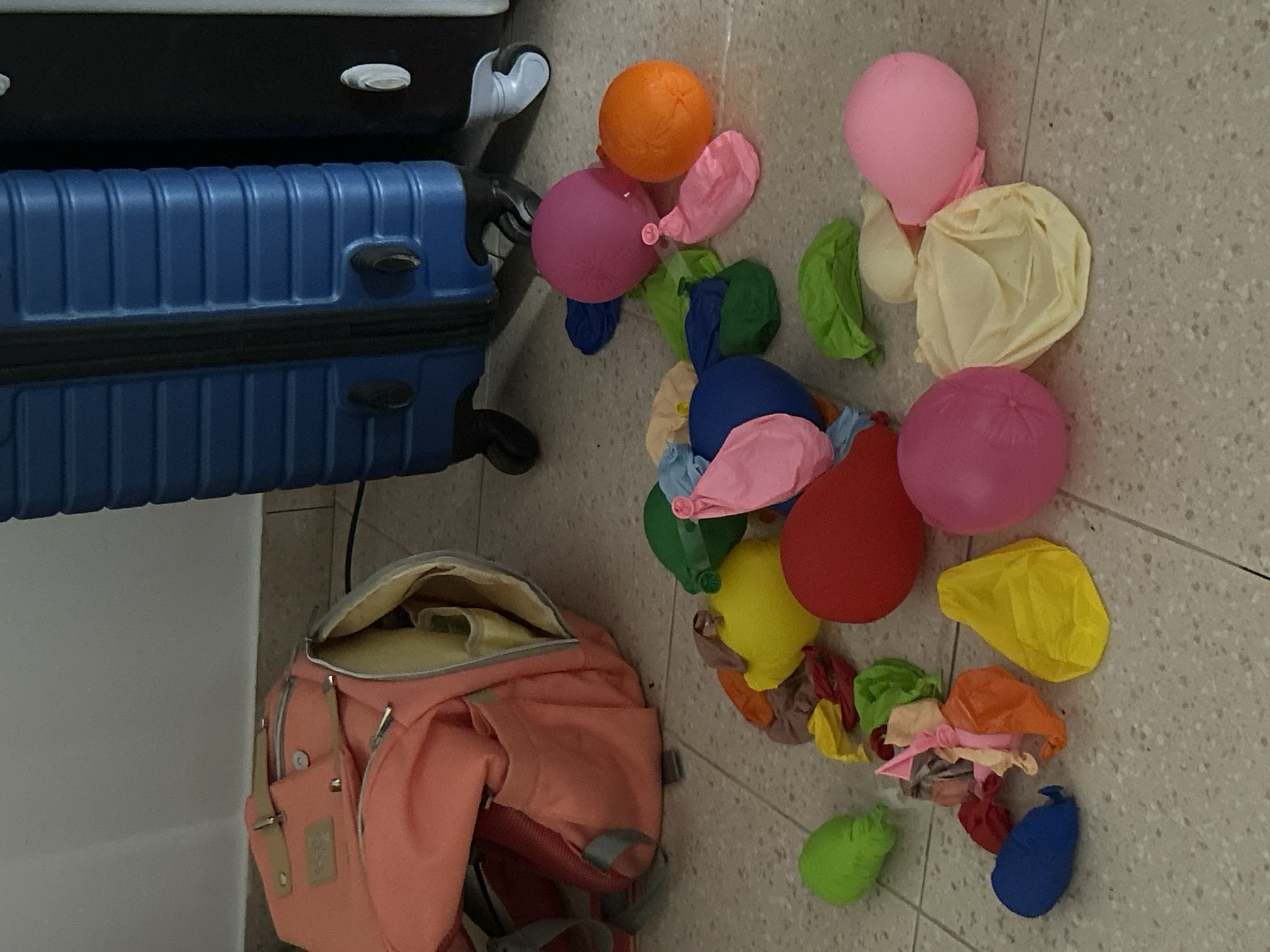 The scene is a tiled floor in an indoor setting, possibly a residence or public space in Câmara de Lobos, Portugal. A blue suitcase rests next to a black suitcase in the upper-left of the image. An open coral backpack is placed next to the blue suitcase. Several deflated balloons of various colors, including orange, pink, yellow, green, blue, and red, are scattered on the floor to the right of the suitcases and backpack. There are no people visible in the image. The environment appears clean and sterile, lacking any organic matter.