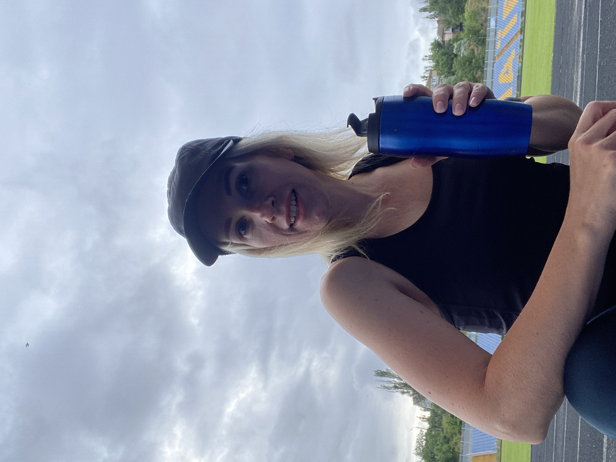 A blonde woman wearing a black tank top and a dark baseball cap is sitting on a track with a blue water bottle in her right hand. She is looking at the camera with a slight smile. In the background, there are stadium bleachers with blue and yellow seats, a green field, and trees on a hillside under a cloudy sky. The scene is set at an outdoor sports stadium.