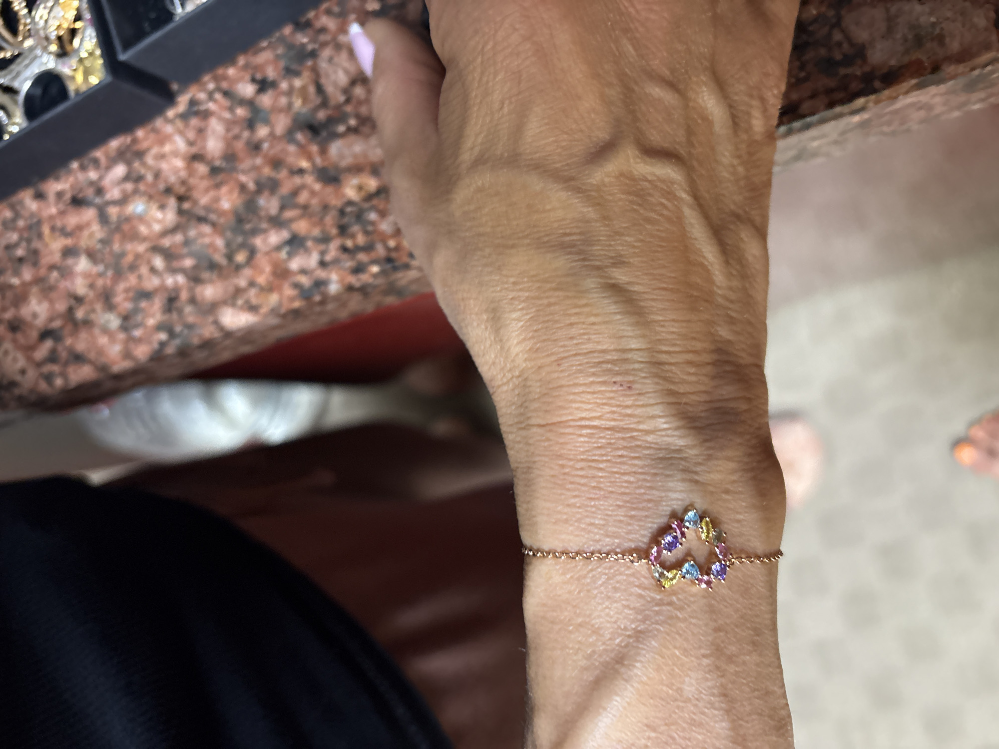 The image captures a close-up of a person's right arm and wrist, featuring a delicate bracelet. The main subject is a light-skinned arm, showing visible veins and skin texture, suggesting an adult individual. The hand is partially visible on the left, with manicured nails painted in a light pink or white French tip style.
A thin, rose gold or copper-toned chain bracelet is worn on the wrist. Its central element is an open heart-shaped charm adorned with multiple small, faceted gemstones in various colors, including light blue, purple, yellow, and pink/orange. The stones are irregularly placed around the heart outline, giving it a vibrant, scattered appearance.
In the upper left background, a dark, possibly black, rectangular object resembling a jewelry box or tray is partially visible, containing what appears to be other metallic jewelry pieces. Beneath this, a reddish-brown and black speckled surface, likely granite or a similar stone countertop, extends across the upper and middle left of the frame. The rest of the background is blurred, showing light-colored surfaces and what might be parts of a person's feet or light-colored footwear on the far right, suggesting an indoor setting.
The image appears to be taken in daylight, though no specific time of day can be determined. There are no discernible emotions or visible text. Based on the provided context, the scene is located in Sofia, Bulgaria.