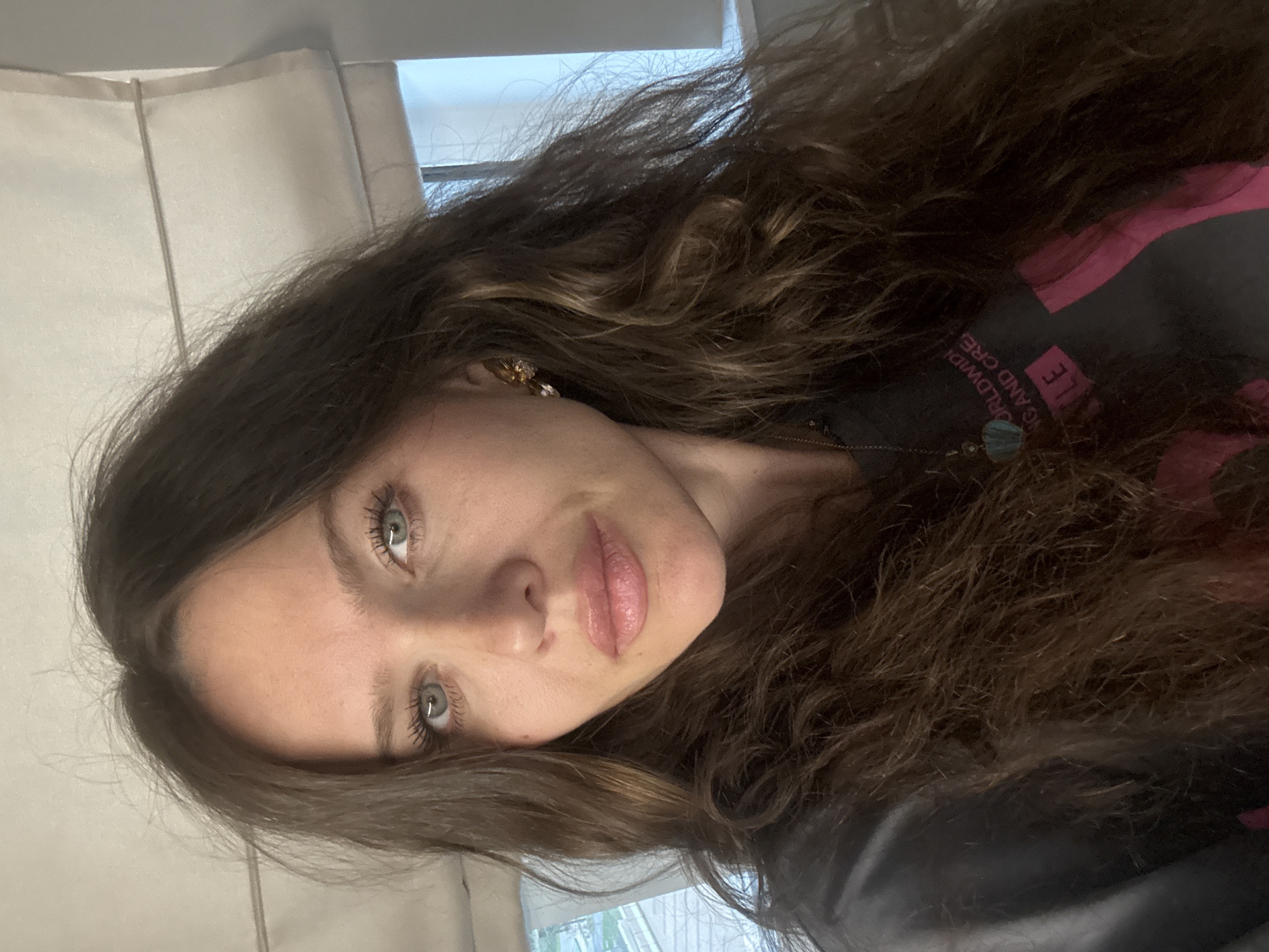 A woman with long, wavy brown hair and light eyes is posing for a selfie. She is wearing a black shirt with pink writing. There is a window and a curtain visible in the background.
There is no information to suggest the location is Khimki, Russia.