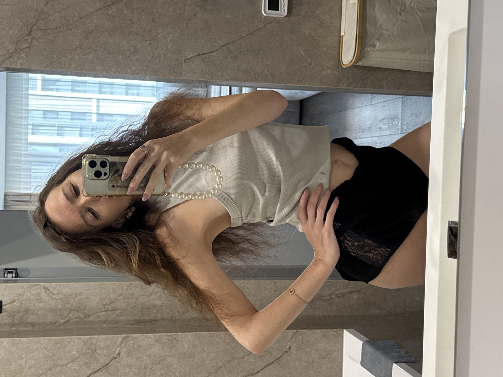 A woman is taking a selfie in a bathroom. She is wearing a light-colored tank top with a pearl necklace and black shorts with lace trim. Her long brown hair is loose. She is holding a smartphone up to her face with her left hand, and her right hand is on her stomach. A gold bracelet is visible on her right wrist. The bathroom has marble walls and a mirror. A high-rise building is visible through a window in the background. The building's location suggests it is likely in Khimki, Russia.