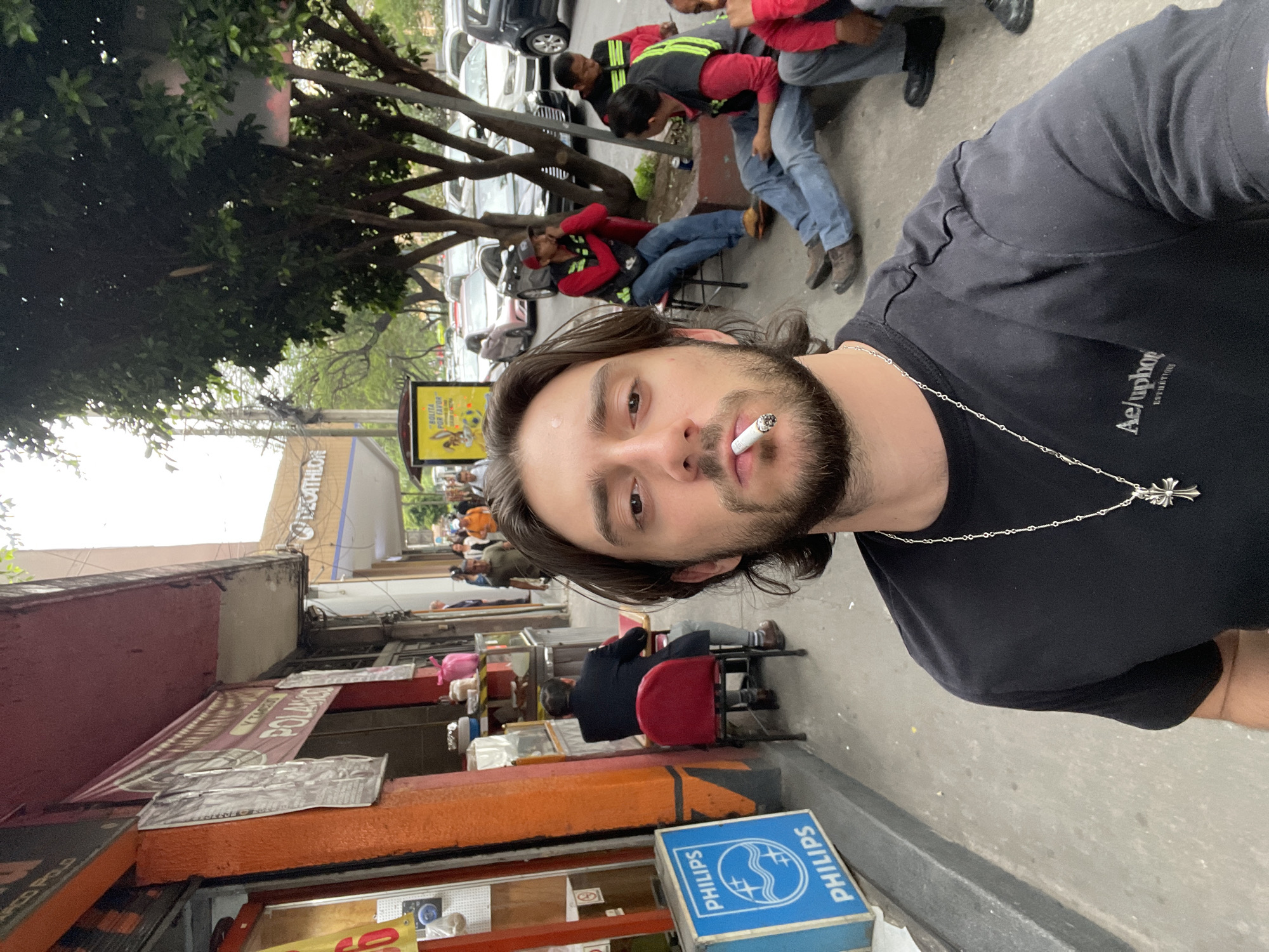 The image is a selfie taken by a man with medium-length brown hair, a beard, and a lit cigarette in his mouth. He is wearing a black T-shirt and a silver necklace with a cross pendant. The background shows a busy street scene. In the immediate background, there are people sitting on chairs along the sidewalk, and storefronts with signs are visible. Further back, there are trees, parked cars, and more people. A yellow sign with advertising is also seen. The location appears to be a street in Mexico City, Mexico. There are men in red shirts and vests leaning around what appears to be a parked car.