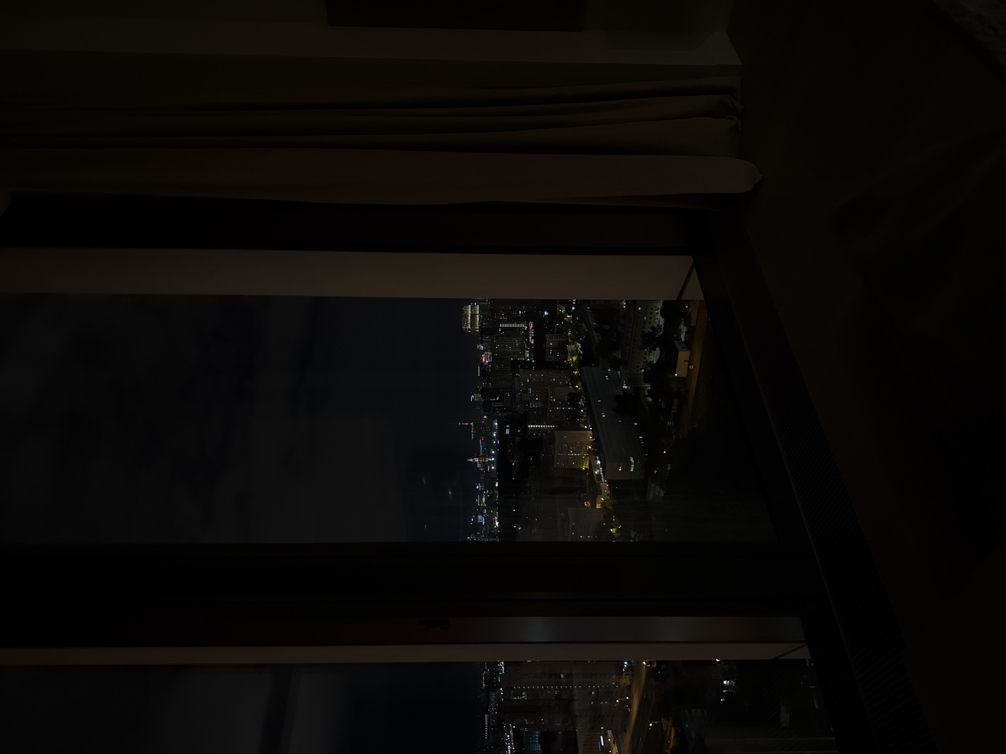A darkened interior view through a window, showcasing a cityscape at night. The city, characterized by scattered lights, consists of high-rise buildings and streetlights. The window frame is dark, contrasting with the dimly lit urban landscape. The immediate foreground is in deep shadow, suggesting an indoor setting. There is no evidence to support the location being in Khimki, Russia. The scene features no people or actions, focusing on the built environment at night.