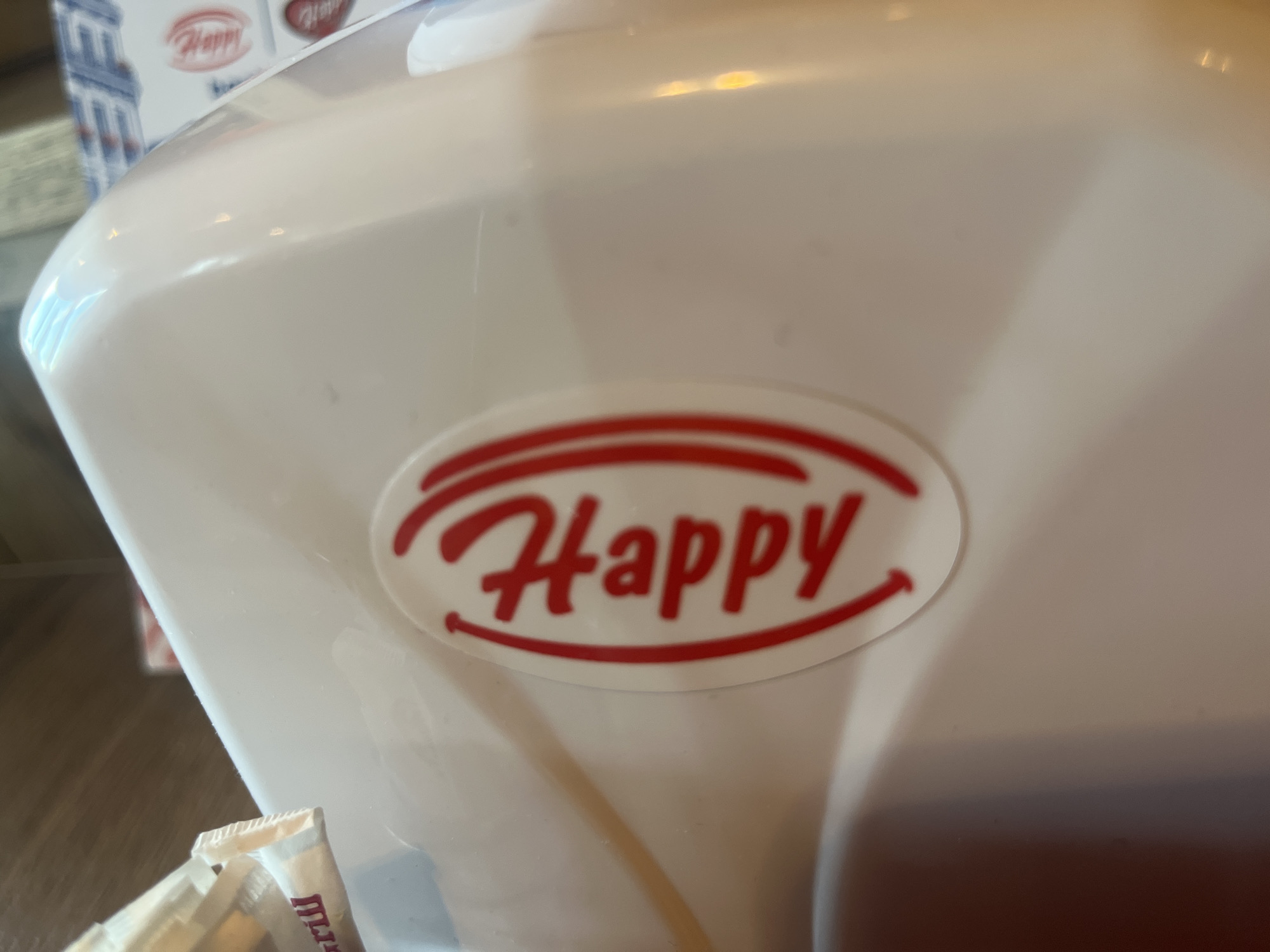 The image displays a close-up of a white, glossy dispenser or container, likely made of plastic, in an indoor setting. The prominent feature is a red logo centrally placed on the dispenser, which spells out "Happy" in a stylized, cursive font. This text is enclosed within a red oval outline, with the bottom curve of the oval subtly shaped like a smiling mouth. The dispenser's surface exhibits reflections from ambient light sources.
In the blurred background, another instance of the same "Happy" logo is partially visible on what appears to be a box or packaging. To the lower left of the foreground, a corner of a small, light-colored packet, possibly containing a condiment like sugar or creamer, is visible. It has some indistinct red text on it. The scene suggests a commercial establishment such as a diner, restaurant, or cafeteria. No people are visible, and no specific activity is depicted. The lighting is consistent with an indoor environment.