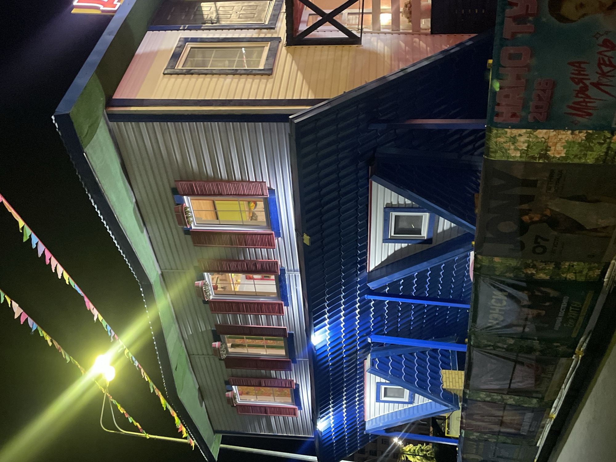 An upside-down house is illuminated at night in Anapa, Russia. The house, painted in muted tones of gray, white, and blue, features red shutters on its windows. In front of the house, a fence is decorated with posters, some of which are partially obscured by the darkness and the structure of the fence. String lights with colorful triangular flags are draped across the top of the scene, casting a festive glow. A street lamp with a bright yellow light is also visible. The overall impression is one of a whimsical attraction at night.