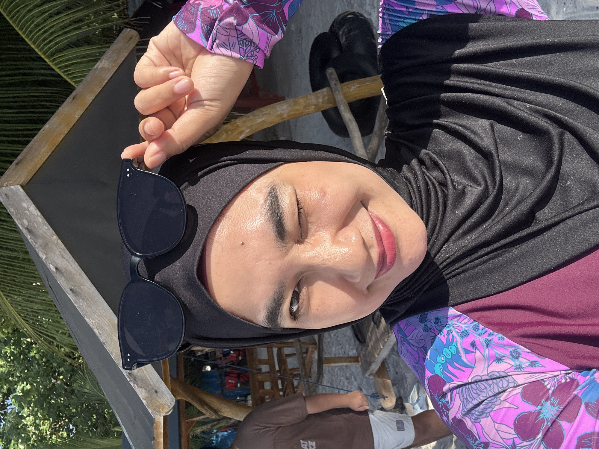 A woman wearing a black hijab and a floral swimsuit is taking a selfie outdoors. She is winking and holding her sunglasses above her head. She is standing on a beach in front of a wooden structure with a thatched roof, possibly a hut or shelter. Palm trees are visible in the background. The woman's face is illuminated by sunlight, and she has makeup on. The environment appears to be tropical.