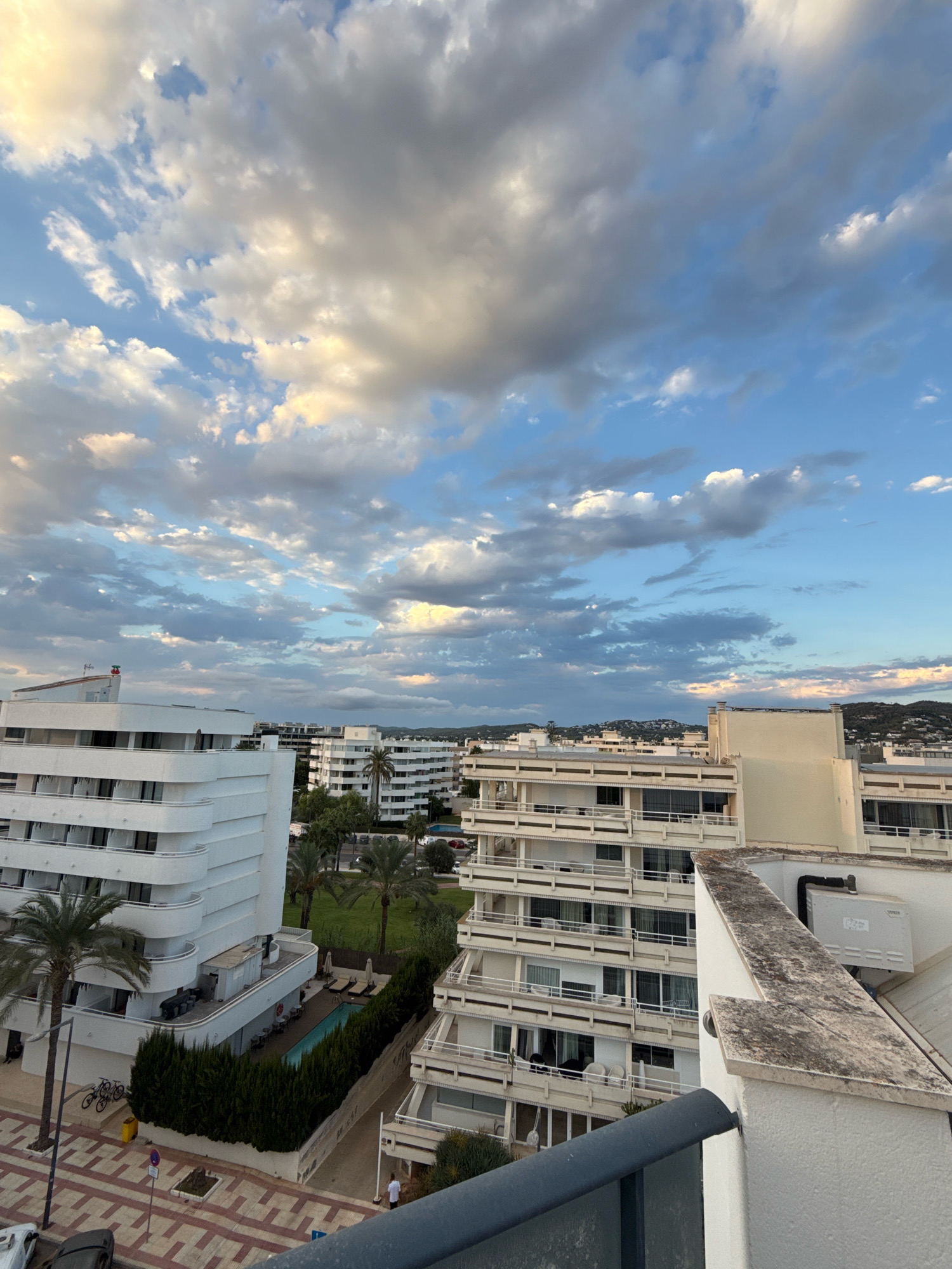 This is a high-angle, wide shot of a residential area in Ibiza, Spain, under a cloudy sky. The buildings are modern, white, and apartment-style, with multiple balconies. In the foreground, the edge of a building with a grey railing is visible. To the left, a white building with curved balconies dominates the frame, with palm trees and bicycles in front. A swimming pool, surrounded by green hedges and lounge chairs, is visible between buildings. Further back, more apartment buildings stretch into the distance, with a hilly landscape in the background. The sky is filled with a mix of white and grey clouds, with patches of blue sky and hints of golden light, suggesting it might be late afternoon or early evening.
