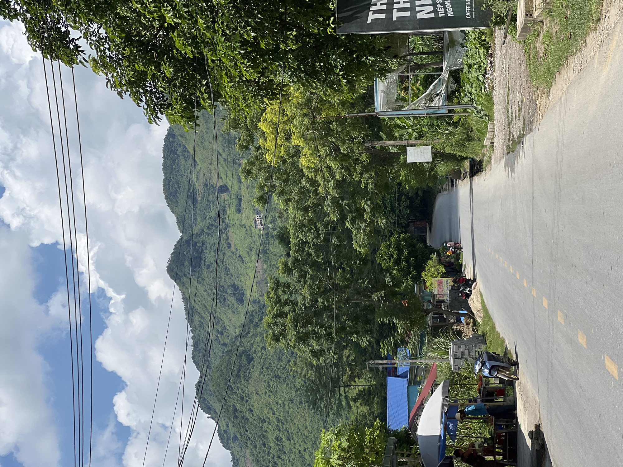 This is a vertical, eye-level, medium shot of a road in Lùng Tám Commune, Vietnam. In the foreground, a paved road with yellow dashed lines runs from the bottom center towards the upper center of the frame. To the left of the road, there is a market stall with a white and blue umbrella, where two people are standing. A motorcycle is parked in front of the stall. Further down the road on the left, there are buildings with blue roofs. On the right side of the road, there is a black sign with white text that appears to be advertising a business. Behind the road and buildings, there are lush green trees and rolling hills covered in vegetation. The sky is blue with scattered white clouds. Power lines are visible in the upper portion of the image, stretching across the frame and in front of the hills. Several other motorcycles are parked or moving along the road.