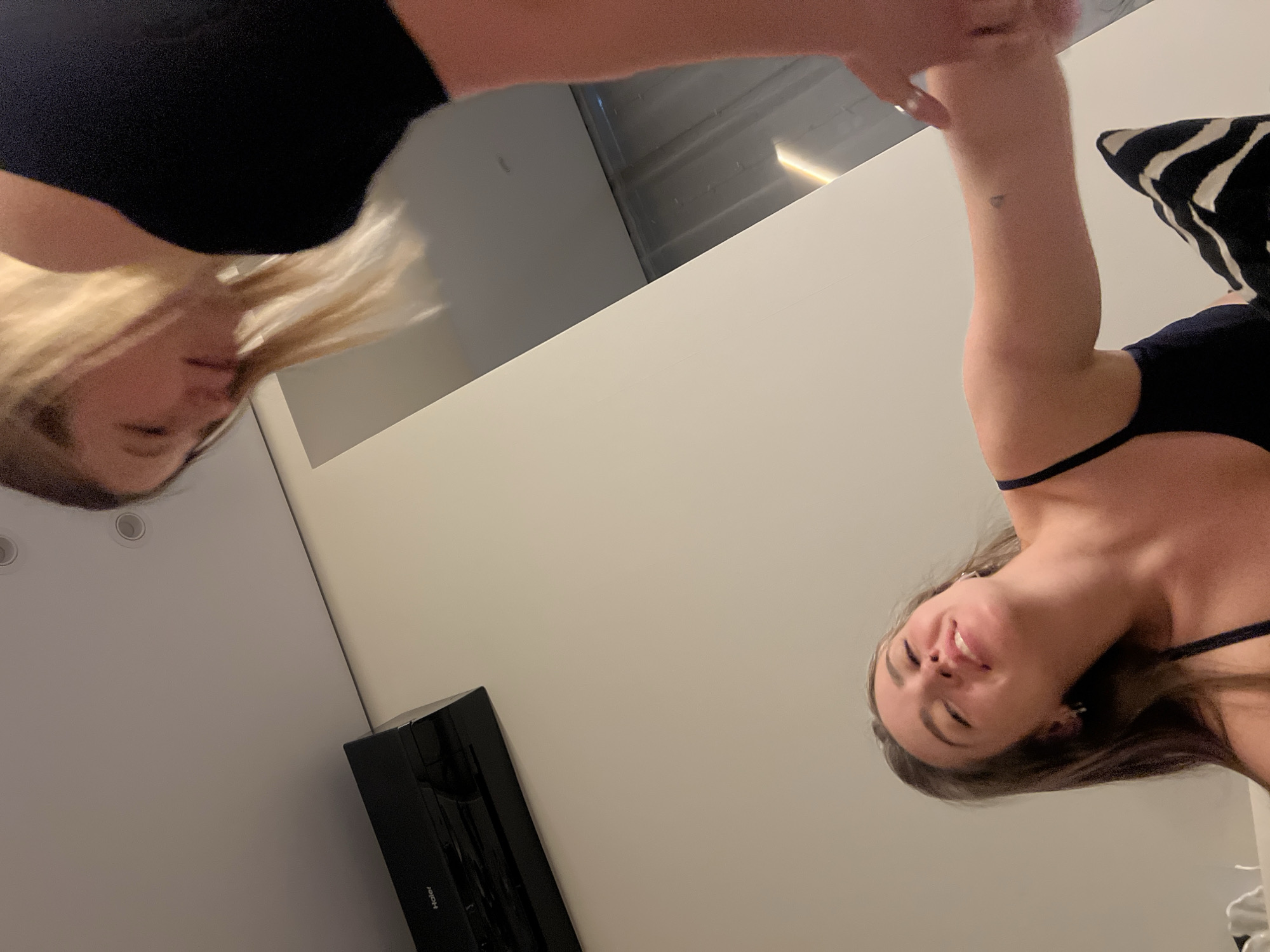 Two women are depicted from a low-angle perspective, possibly looking down. One woman has blonde hair and is wearing a black top. The other woman has brown hair and is wearing a similar black top with spaghetti straps. A zebra-patterned pillow is partially visible. A black object, possibly an electronic device, is situated along the bottom left edge of the frame. The background is composed of light-colored walls and a ceiling with recessed lighting. This image appears to be taken in a domestic setting. The location of Рубцово, Russia is not evident from the image.