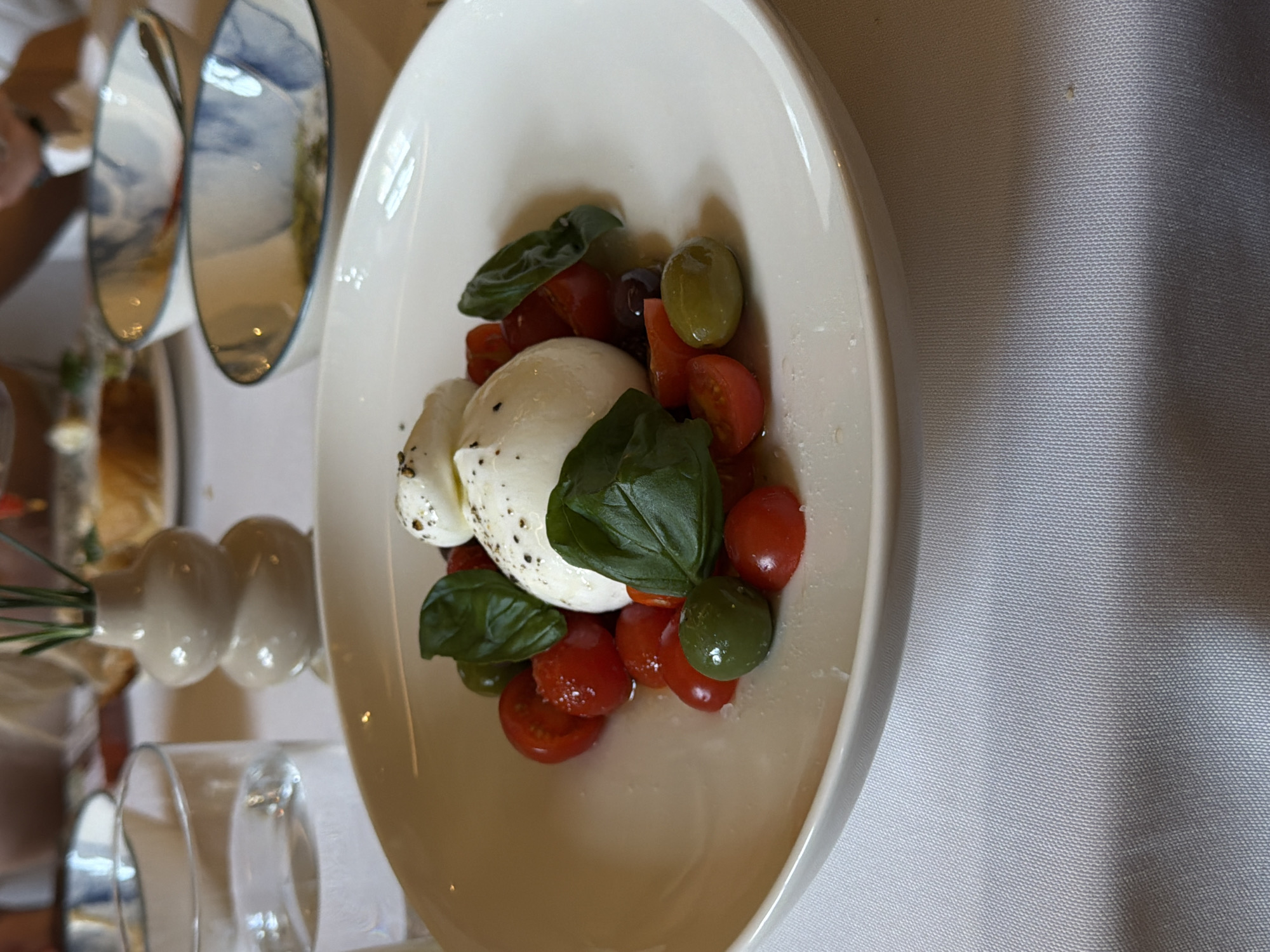 A close-up shot focuses on a white plate of burrata cheese, cherry tomatoes, basil, and olives, presented on a white tablecloth. To the left of the plate, a glass of water is visible. In the background, a small decorative vase with greenery and a blurred image of a dining table with food and bowls complete the scene. The setting is a restaurant in Kyiv, Ukraine.