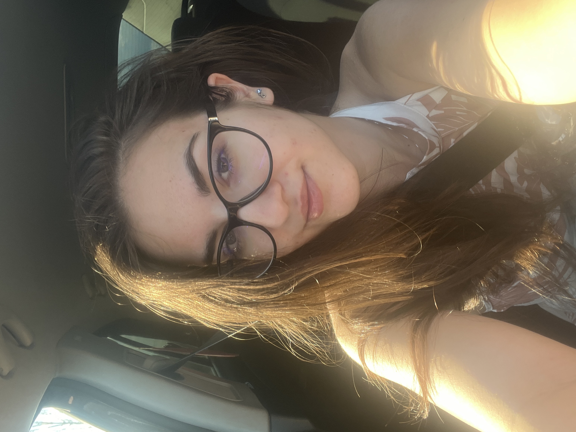 A woman with long brown hair, wearing black-rimmed glasses and a seatbelt, sits in a car. The photo is taken from a slightly elevated angle. The lighting is bright, potentially sunlight, with some glare on the hair and skin. The background includes the car's interior and a glimpse of the outside environment. The location is not discernible from the image. No identifiable landmarks or specific features indicate the location is Sofia, Bulgaria.