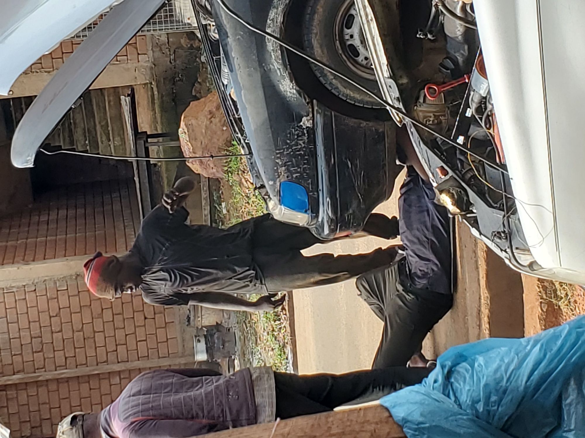 In Bukuru, Nigeria, two men are observed working on a vehicle with its hood open. One man, wearing a red and brown beanie and a black t-shirt, stands next to the vehicle and appears to be gesturing or reaching into the engine bay. Another man is lying on the ground beneath the vehicle, his legs extended, suggesting he is performing maintenance from underneath. A third individual is partially visible on the left side of the frame, standing and facing away from the camera. The scene is outdoors, with a brick wall and stairs visible in the background. The ground appears to be dirt or asphalt. Two cars are present, one black and one white, both with their hoods open, indicating repair work is in progress. The overall impression is a street-side repair scene in a Nigerian urban or semi-urban environment.