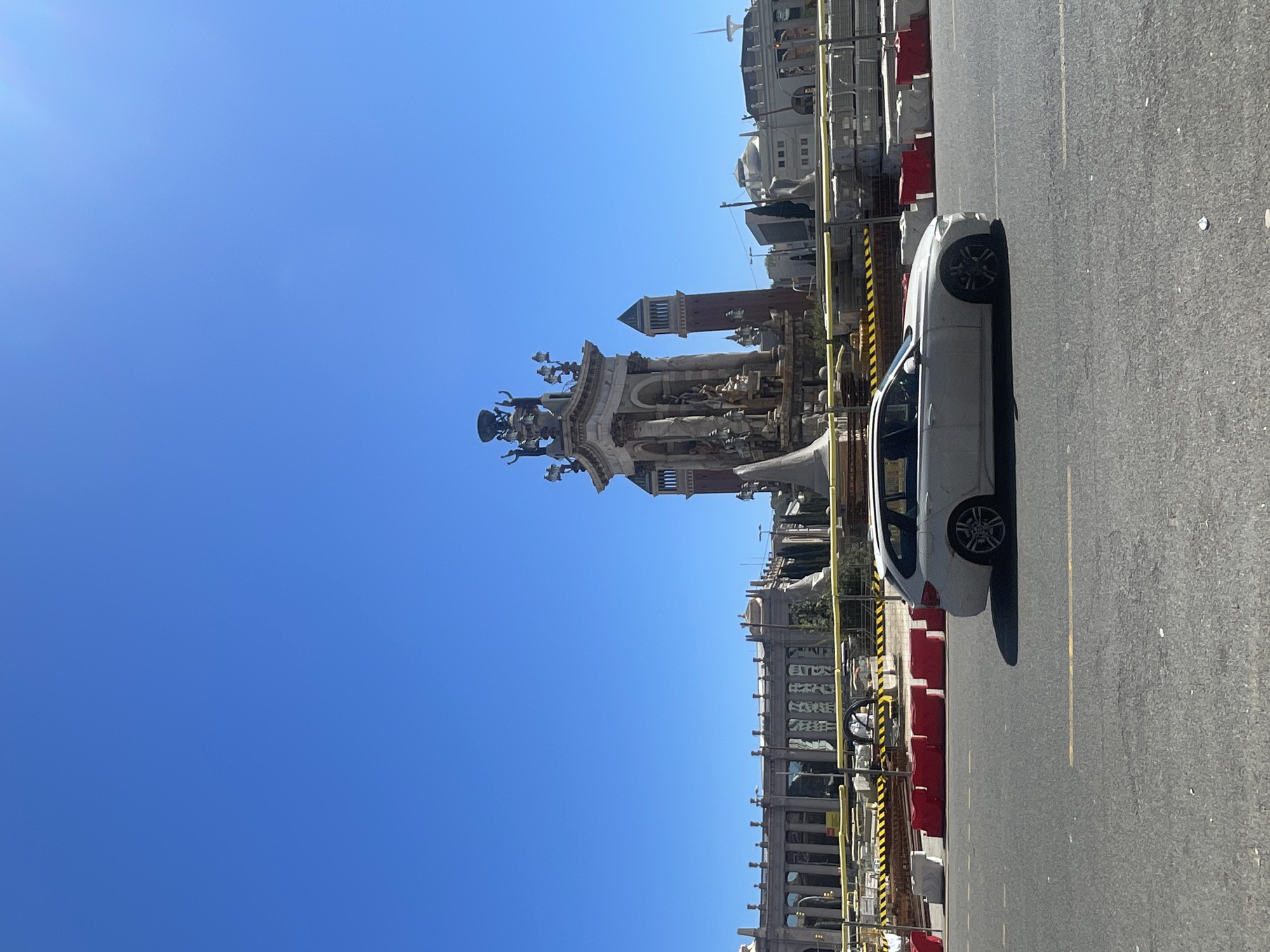 The image captures a sunny day in Barcelona, Spain, featuring the Magic Fountain of Montjuïc and a section of the Venetian Towers in the background, set against a clear blue sky. A white car is parked on the right side of the frame, along a road or street. Construction barriers, marked in red and yellow, stand along the roadside. No people are visible in the frame.
