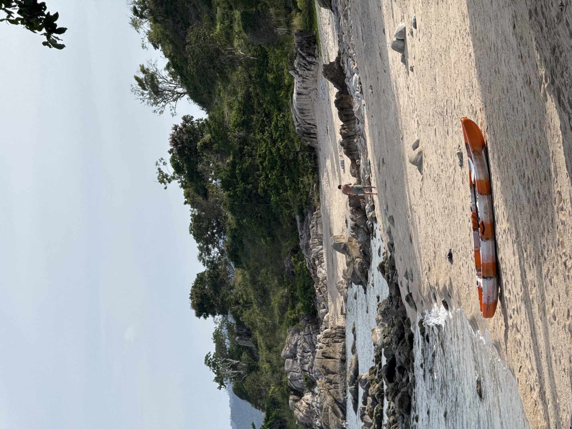 A bright, slightly overcast day at a rocky beach in Koh Samui, Thailand. The foreground features a sandy beach with a two-person kayak resting on its side, painted in orange and white. Waves gently lap at the shore. In the middle ground, a lone man in shorts walks away from the camera towards a rocky outcrop. The background is dominated by a lush green hillside covered in trees and dotted with large rock formations. The sea is visible to the left of the frame, with more rocks emerging from the water.