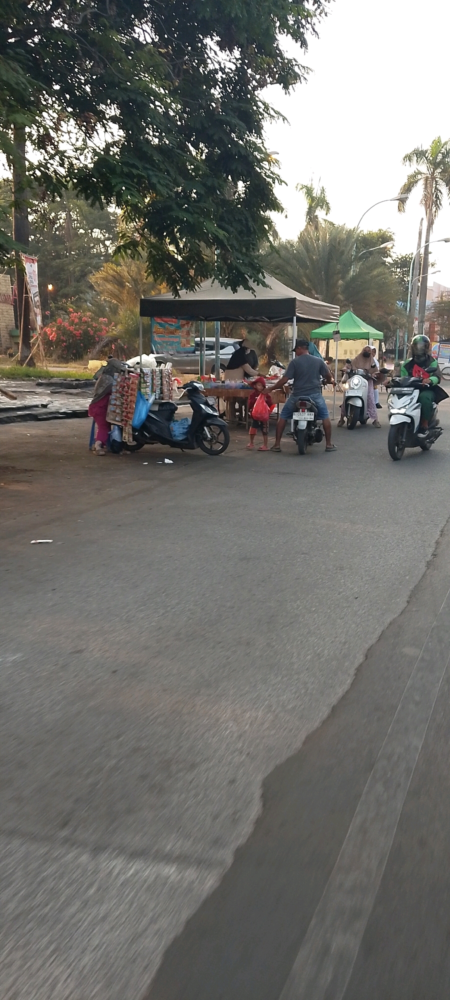 In Karangsatria, Indonesia, on a paved road, a street vendor displays an array of bagged goods from a scooter. To the left, two individuals, one wearing a pink headscarf and pink clothing, stand near the scooter. A black scooter is parked on the left side of the scene. In the background, a vendor operates from under a dark tent, with a variety of items on display. Further back, a white car is visible, and beyond that, a green tent. Two individuals are on a white scooter, and another person is on a white scooter in the far right, wearing a helmet. A child in red shorts and a red shirt stands near the vendor under the dark tent, holding a plastic bag. A man in a grey t-shirt and blue shorts is on a white scooter, facing away from the camera. The environment suggests a marketplace or roadside stall area with trees and palm trees visible in the background. The road has a distinct texture, and there are some debris items on the ground.