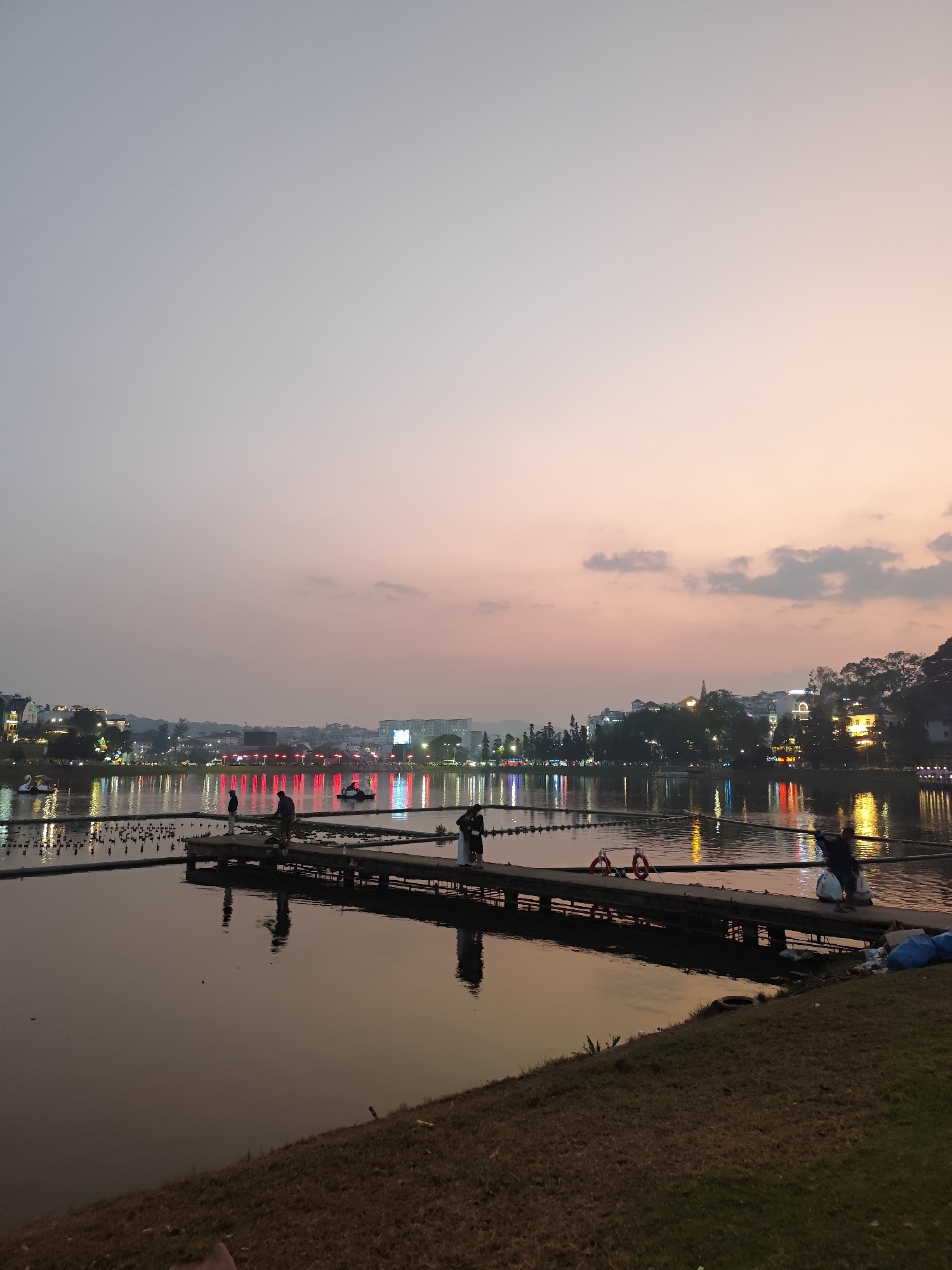The image depicts an outdoor twilight scene at Xuan Huong Lake in Da Lat, Vietnam.
In the foreground, a sloping grassy bank occupies the lower right, with several blue bags and a dark tire visible near the water's edge. A dark wooden pier extends from the right foreground into the lake, featuring two red-and-white life preservers hanging from its side. Four individuals are present on or near the pier: one sits on the far right edge, two stand further along the pier facing the water, and one is partially visible on the far left.
The mid-ground comprises the lake's surface, reflecting the illuminated city lights. Scattered water lilies are visible near the pier. A horizontal line of small, dark, buoyant objects, possibly buoys, stretches across the water. Several swan-shaped pedal boats and other small vessels are dispersed on the lake.
The background showcases the far bank of the lake, lined with buildings illuminated by various colored lights (predominantly red, white, yellow, and blue), which create distinct reflections on the water. Trees are interspersed among the structures.
The sky transitions from a light grey-blue at the top to a soft orange-pink near the horizon, with scattered faint grey clouds. The overall lighting conditions indicate dusk or early evening.