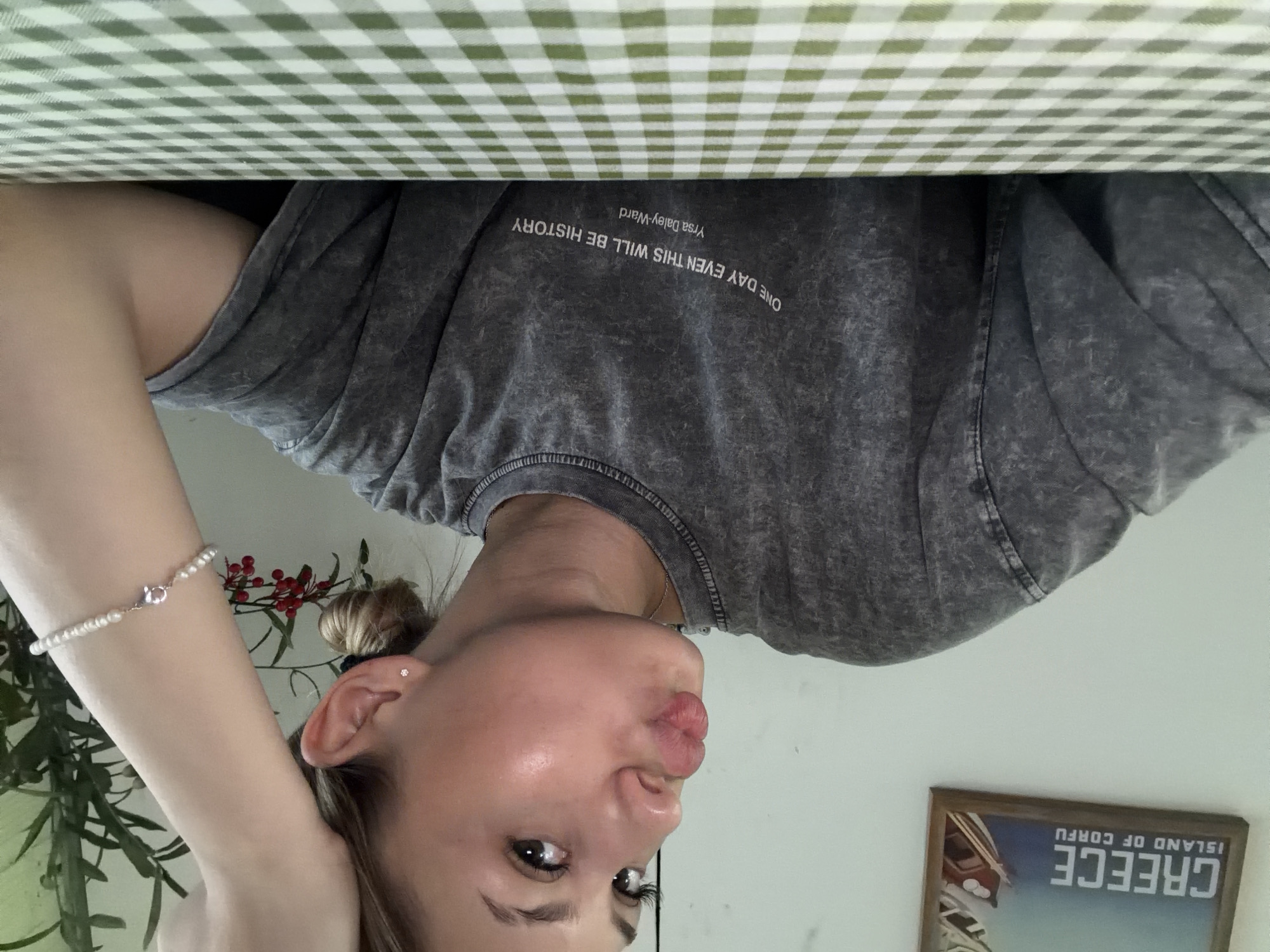 The image depicts a young Caucasian woman with blonde hair pulled back in a bun. She is wearing a grey t-shirt with the text "ONE DAY EVEN THIS WILL BE HISTORY Yrsa Daley-Ward" printed on it. She is looking to her left and making a kissing gesture with her lips. She has a pearl bracelet on her right wrist and a small stud earring in her left ear. In the background, a framed picture of Greece is visible on the wall, and a green potted plant with red berries is also present. The woman is seated at a table covered with a green and white checkered tablecloth. The image is taken indoors, likely in a residential setting, and does not contain any elements that would indicate a specific location in Da Nang, Vietnam.