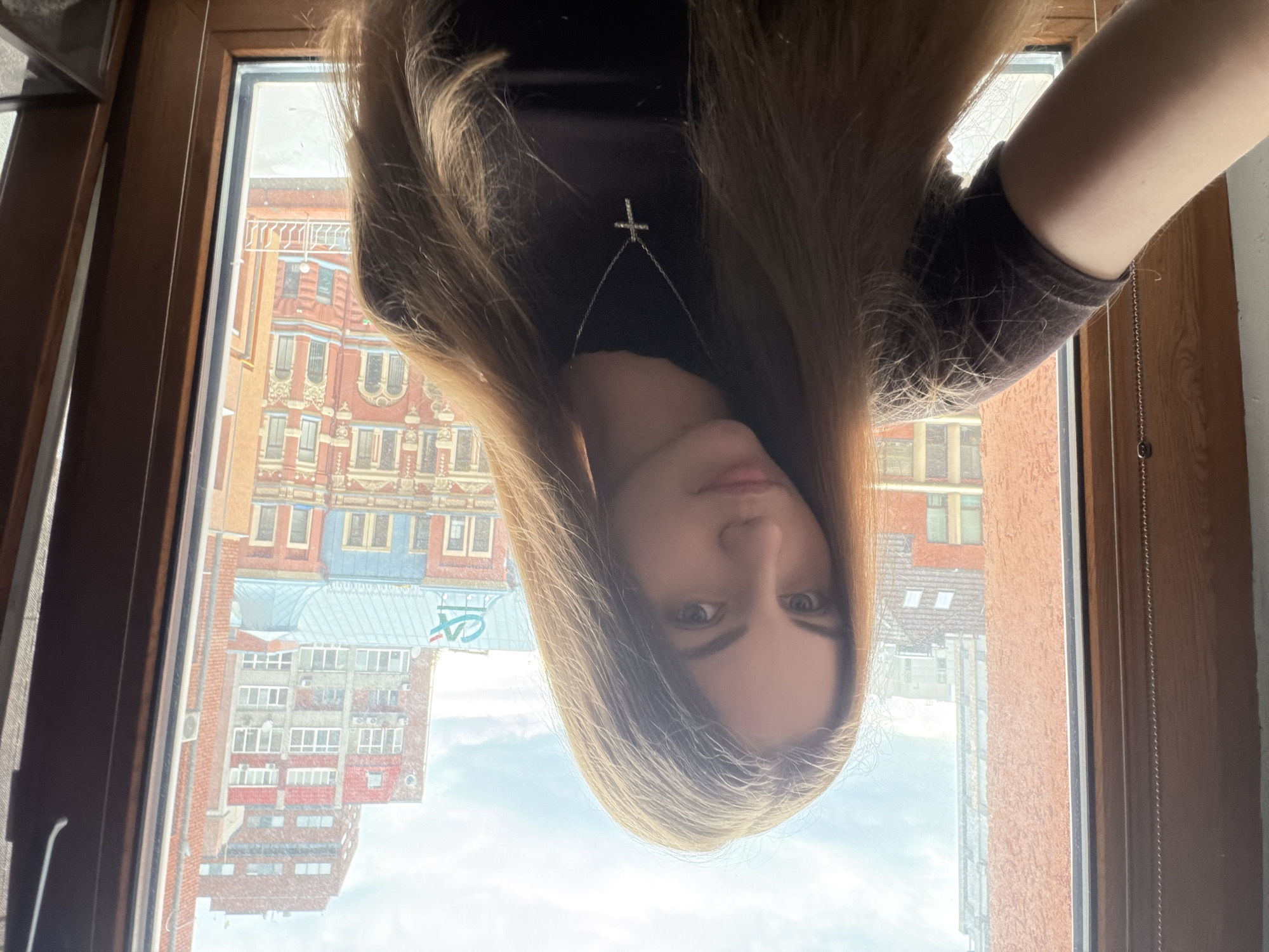 A young woman with long, auburn hair takes a selfie in front of a window with a view of buildings. She is wearing a dark t-shirt and a silver cross necklace. The background shows urban architecture with red brick buildings and a cloudy sky. The location appears to be in the city of Dnipro, Ukraine, given the architectural style and the presence of a "CA" logo on one of the buildings, which is associated with Crédit Agricole, a French bank with a presence in Ukraine.