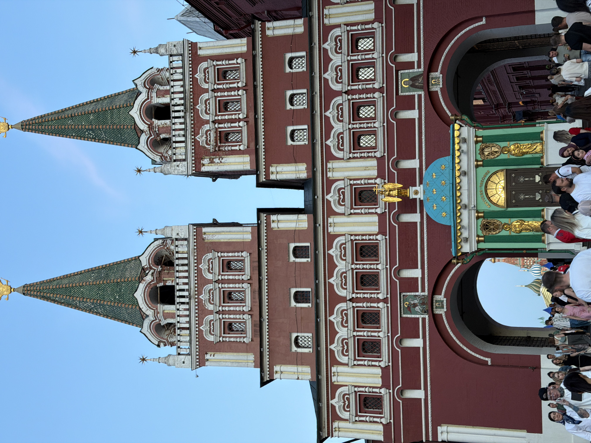 This image depicts the Iberian Gate and Chapel, also known as the Resurrection Gate, located at the entrance to Red Square in Moscow, Russia. The scene captures a bustling crowd of individuals passing through and around the historical structure under a clear blue sky.
The central architectural feature consists of two symmetrical towers constructed from red brick with contrasting white decorative elements. Each tower features multiple levels of arched windows with grilles, varying in size and arrangement. The upper section of each tower includes an open-arch balcony area with white balustrades. Both towers are capped with steep, green-tiled conical roofs, each surmounted by a golden double-headed eagle emblem.
Positioned between the two large archways at the base of the gate is the Iberian Chapel. This ornate, small structure has a bright teal-blue domed roof adorned with golden stars. Its facade is green with gold trim and features two golden statues flanking a central entrance with a semicircular window above the doors. A golden angelic figure holding a cross is mounted on a pedestal above the chapel's entrance. Icons are present on the gate above each main archway and within the architectural details.
Numerous people populate the foreground and background. They are dressed in casual clothing suitable for warm weather, indicating a diverse mix of ages and apparent activities such as walking, observing, and conversing. Some individuals are clearly visible in the foreground, while others are a blur of motion through the archways, suggesting a constant flow of foot traffic. Through the archways, additional structures with distinctive colorful domes, likely St. Basil's Cathedral, are partially visible in the distance, further confirming the location within Moscow's Red Square area. The lighting suggests daytime conditions, with soft shadows and clear visibility.
