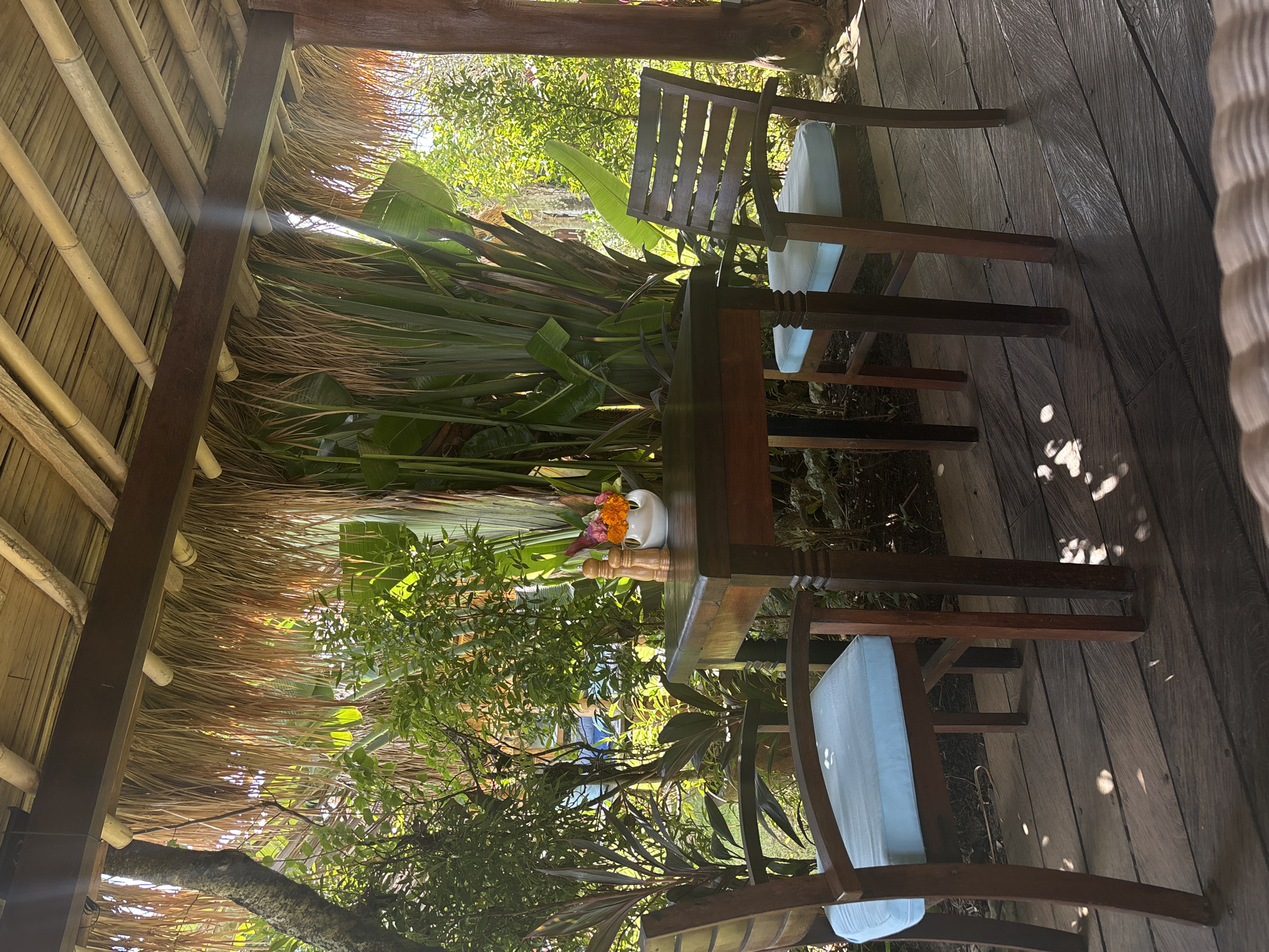 The image depicts a covered outdoor dining area, likely a part of a resort or villa, situated in Kuta Selatan, Indonesia. The structure has a bamboo and thatched roof, suggesting a tropical or Balinese design. A small wooden table is centered in the scene, adorned with a white vase holding a bouquet of orange and purple flowers. Two wooden chairs with pale blue cushions flank the table. The background features dense green tropical foliage. The flooring appears to be made of dark-stained wooden planks. The scene is illuminated by natural sunlight filtering through the vegetation and roof.