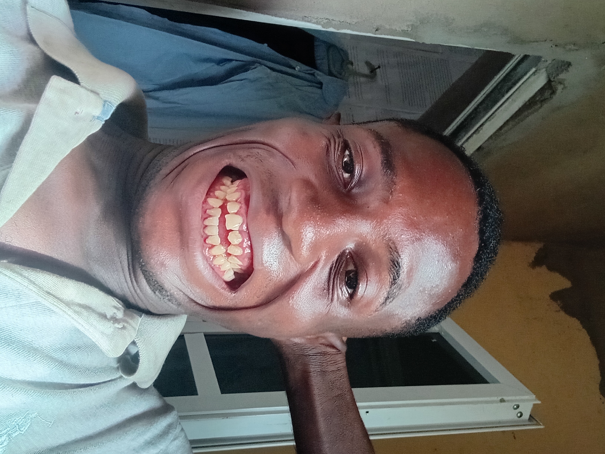 A portrait of a man with dark skin and short, dark hair. He is grinning, revealing his teeth. He is wearing a light-colored shirt. His arm is raised to his head. In the background, there is a window frame and what appears to be a blue piece of clothing. The location in the city of Akwete, Nigeria is unspecified.
