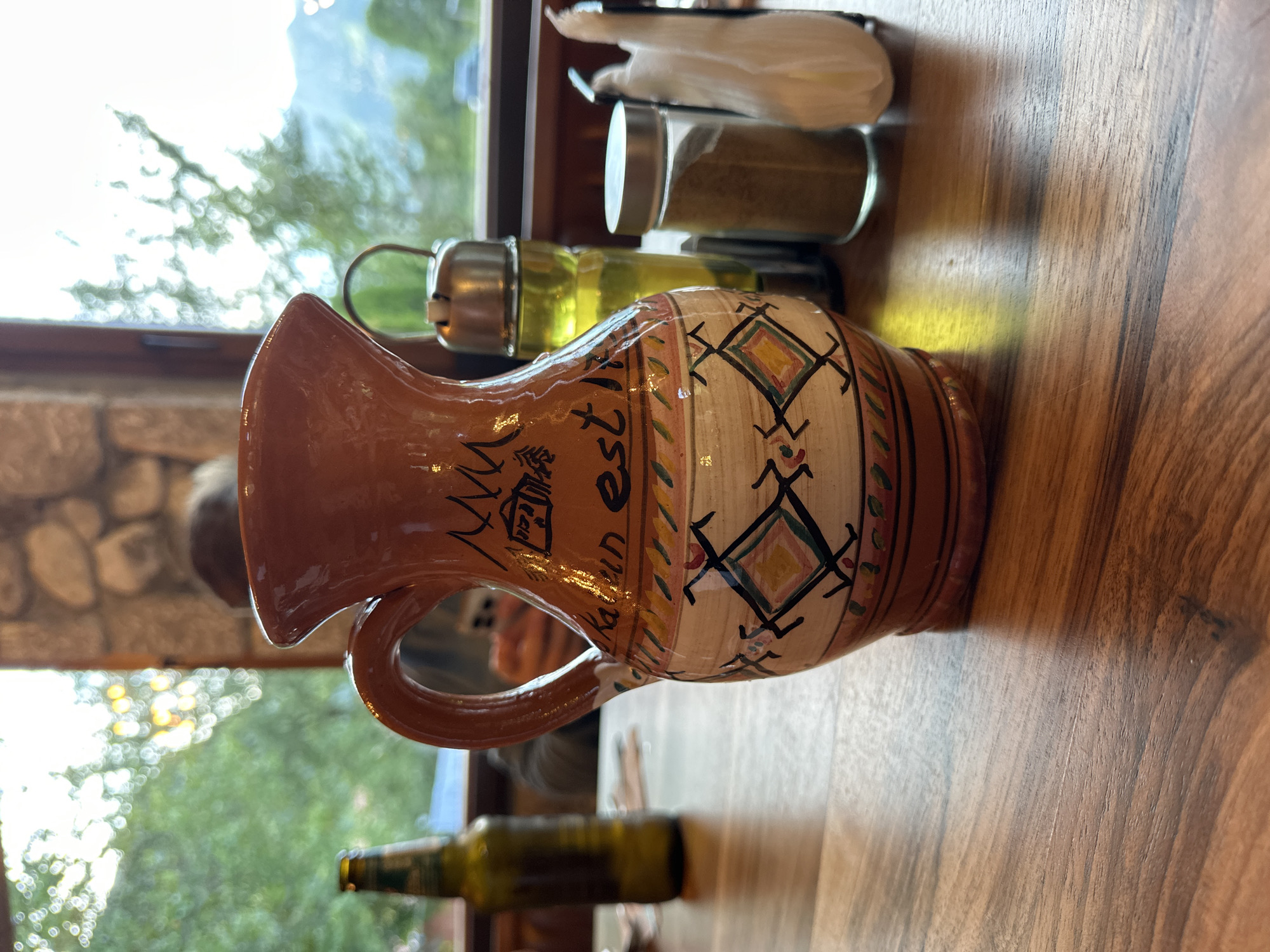 A medium shot captures a decorative, earthy-toned ceramic pitcher on a wooden table. The pitcher is embellished with hand-painted geometric patterns in various colors, including diamond shapes with yellow, blue, and red accents, and abstract designs in black. A building and some wavy lines are depicted in black on the neck of the pitcher.
To the left of the pitcher is a dark green glass bottle with a label that is partially visible. Behind the pitcher, a clear glass bottle filled with a yellow liquid, likely olive oil, stands next to a small glass jar containing a brown granular substance, possibly a spice. A white folded napkin is to the right of the spice jar.
The background reveals a softly blurred view through a window, showcasing lush green trees and a hazy mountainous landscape under a bright sky. The stonework of a building is visible on the upper left. The scene suggests an indoor setting, likely a restaurant or cafe, in Gusinje, Montenegro, where the natural beauty of the surroundings is visible through the window. The lighting appears to be natural daylight, creating a warm and inviting atmosphere.