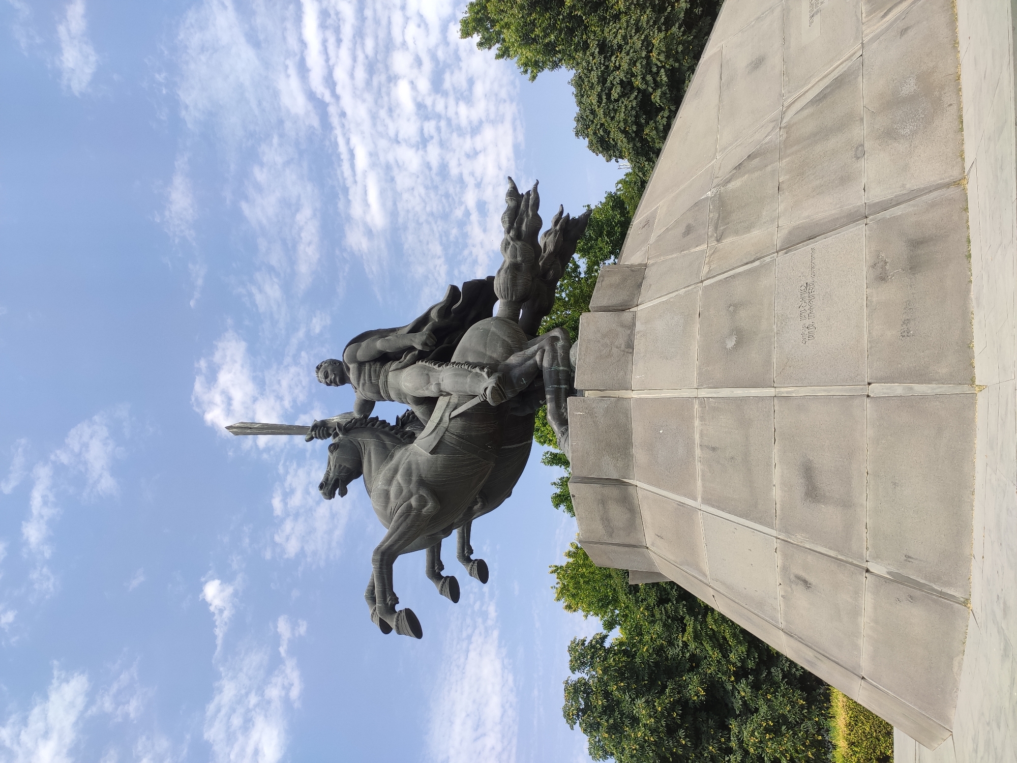 A bronze equestrian statue of a man holding a sword is depicted against a blue sky with white clouds. The statue is positioned on a conical stone monument. The man is muscular and wears a cape. The horse is rearing on its hind legs, with its front legs raised. The monument has a faceted surface, with visible inscription on its side. Green trees are visible in the background. The lighting suggests it is daytime.