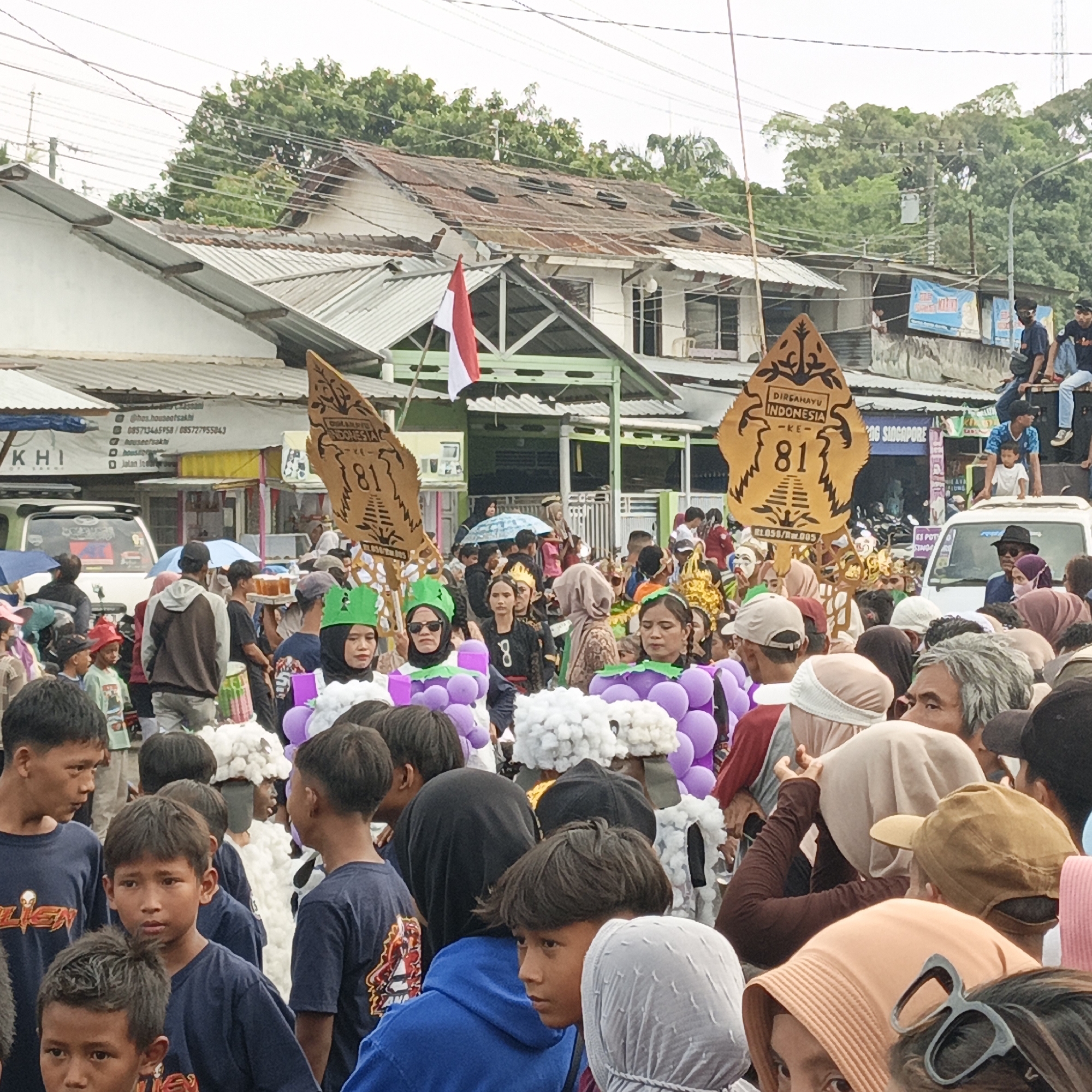 This image captures a lively street scene in Randudongkal, Indonesia, likely during a festive occasion. A crowd of people, including many children and adults, are gathered, some wearing distinctive costumes. In the foreground, several children in dark blue shirts are visible, one looking directly at the camera. Behind them, individuals are adorned with large, purple, grape-like decorations on their heads and shoulders. A person in a white, fluffy costume stands out in the mid-ground. Two large, wooden wayang-shaped props, inscribed with "DIRGAHAYU INDONESIA 81", are prominently displayed, suggesting a celebration of Indonesian Independence Day. An Indonesian flag, red and white, flutters in the background. The environment appears to be an urban setting with buildings, electrical wires, and trees lining the street. A white vehicle is parked in the middle ground. Some people are holding umbrellas, indicating possible light rain or strong sun. The overall atmosphere is one of community celebration and cultural expression.