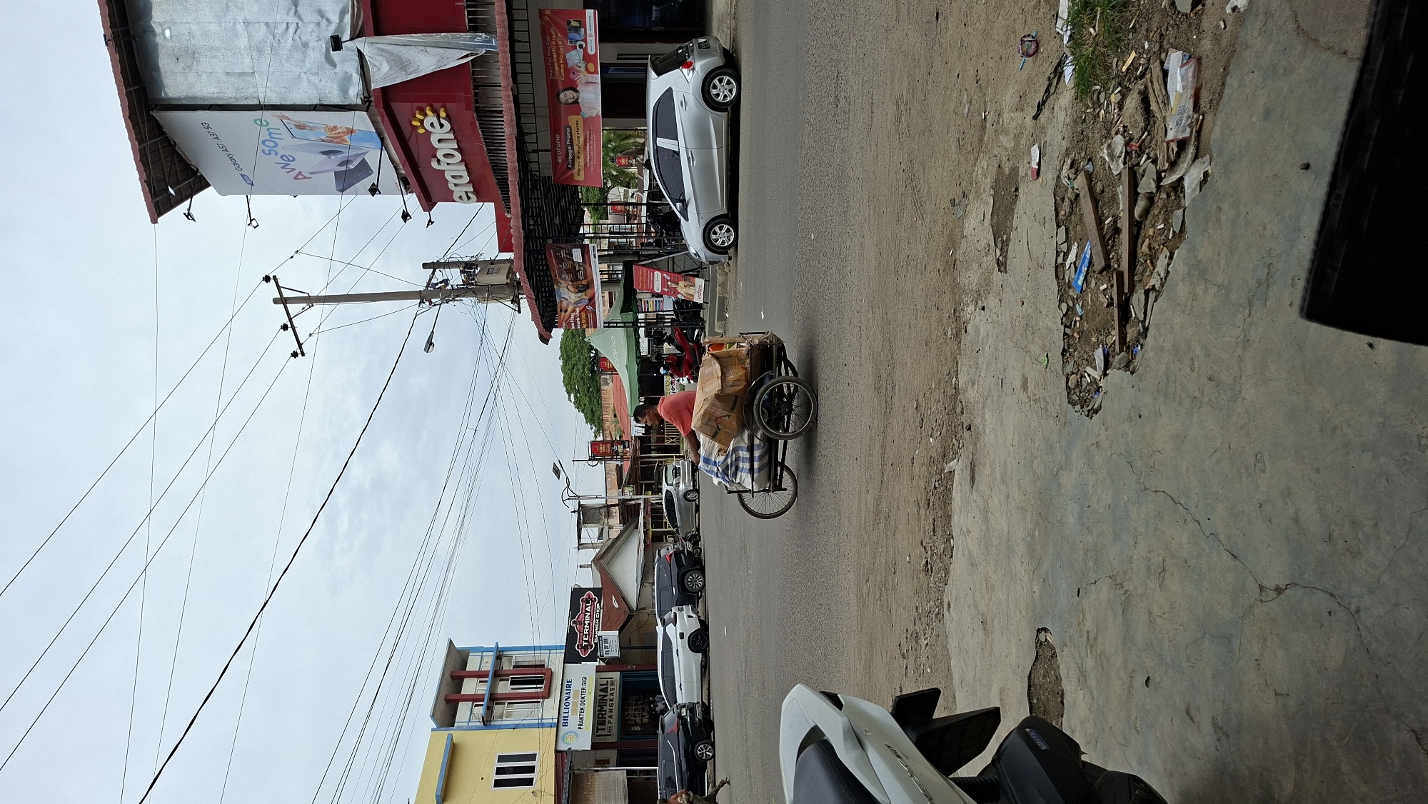 The image depicts a street scene in Lhokseumawe, Indonesia, featuring a man pushing a bicycle-powered cart loaded with goods. The cart is laden with packages, some wrapped in brown cardboard and others in blue and white striped fabric. The man, wearing a red shirt, is actively engaged in moving the cart across the asphalt road.
Several vehicles are present on the road, including a silver car on the right, a white car and two black cars parked further back on the left. In the background, various buildings are visible. To the left, a two-story yellow and blue building with a sign that reads "BILLIONAIRE PRAKTEK DOKTER GIGI TERMINAL PANGKAS" stands out. Adjacent to it is a smaller building with a red roof. On the right side of the road, a red building with a prominent "Erafone" logo is situated. Above the Erafone building, a large billboard advertising "Galaxy A51 A51 5G AWESOME" is displayed.
The environment appears to be a commercial area with shops and businesses lining the street. Overhead, numerous power lines crisscross the sky, supported by utility poles. The ground in the foreground shows a rough, uneven concrete surface with some debris and cracks. The overall atmosphere suggests a typical day in an urban setting, with economic activity and pedestrian and vehicular movement.