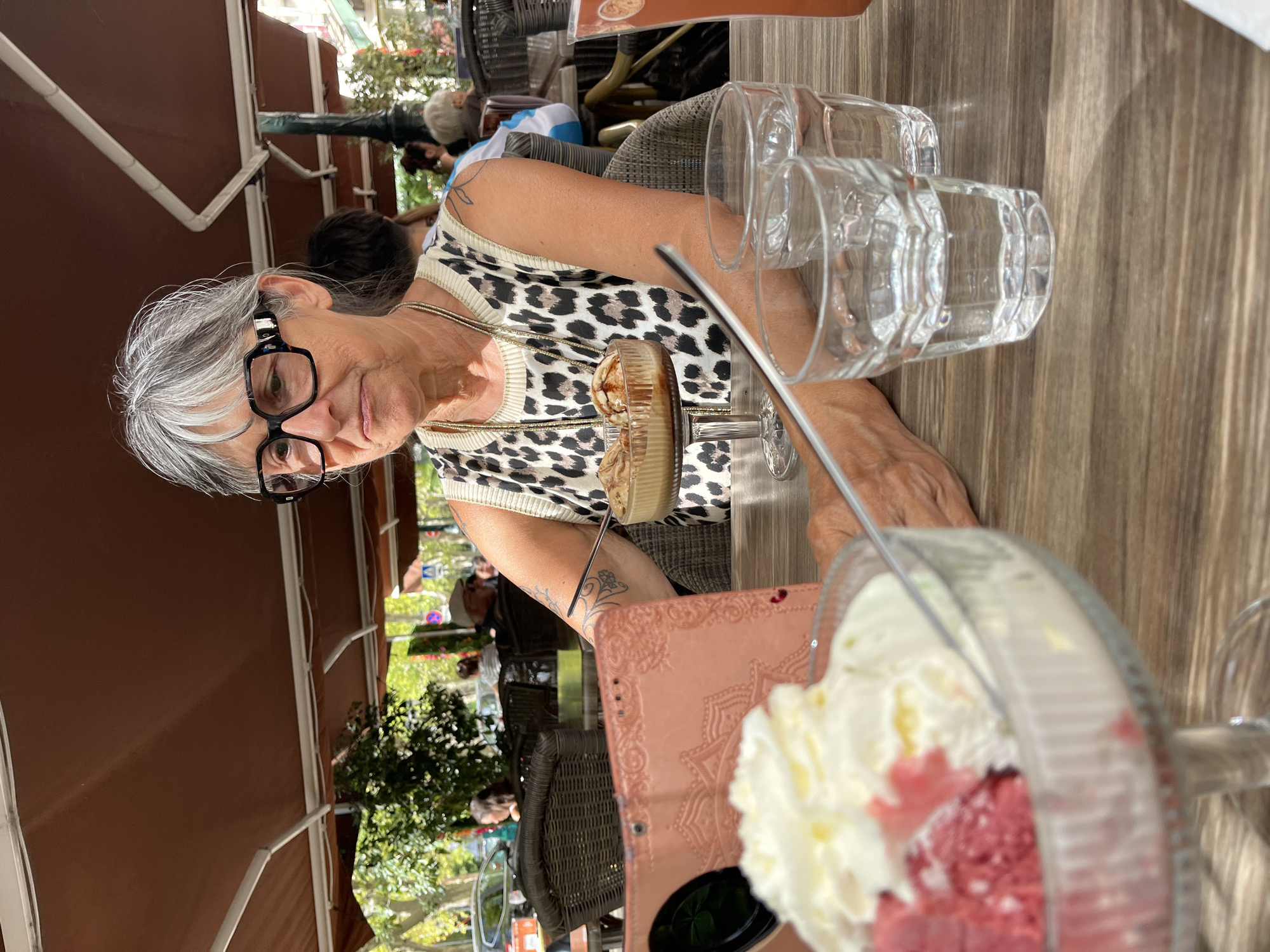 The image depicts an outdoor cafe scene in Montélimar, France, on a sunny day. In the foreground, a woman with gray hair and large black-rimmed glasses is seated at a wooden table. She is wearing a sleeveless top with a leopard print pattern and a gold necklace. In front of her are two glasses of water, and a dessert bowl containing what appears to be ice cream. The woman is looking to her left, and her right arm is resting on the table. A pink tablet is also visible on the table. In the background, other patrons are seated at tables under a brown awning. Trees and buildings are visible in the distance, suggesting an urban or town setting. The overall atmosphere is relaxed and casual.
